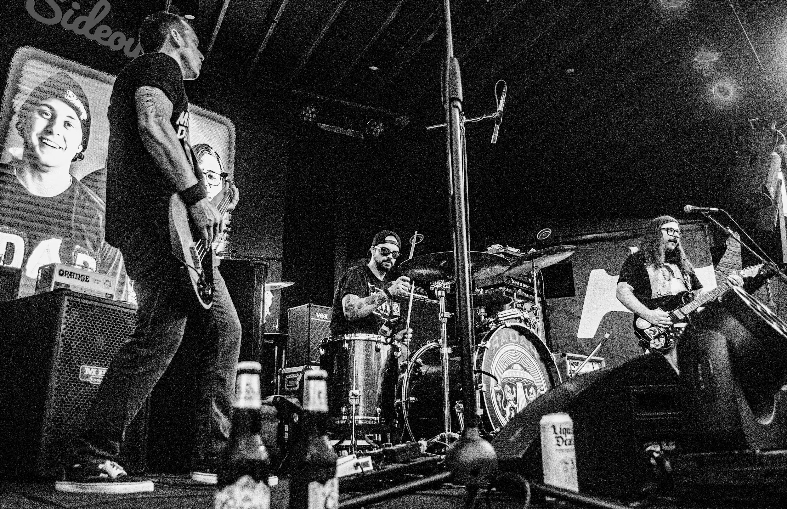 A black and white photo of a live music performance on stage, featuring a group of musicians including a drummer, a guitarist, and a bassist. The background includes posters of smiling individuals and sound equipment.