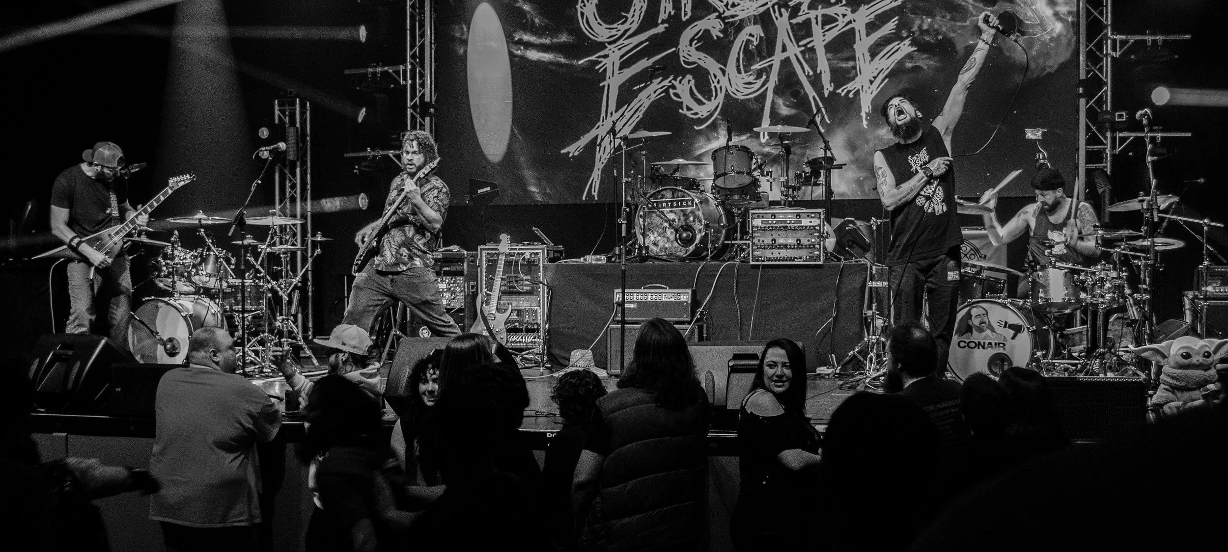 Black and white photo of a live music band performing on stage. Five musicians are seen playing various instruments including guitar, drums, and microphone. The background features a large screen with a design and the word 'ESCAPE'. Audience members 
