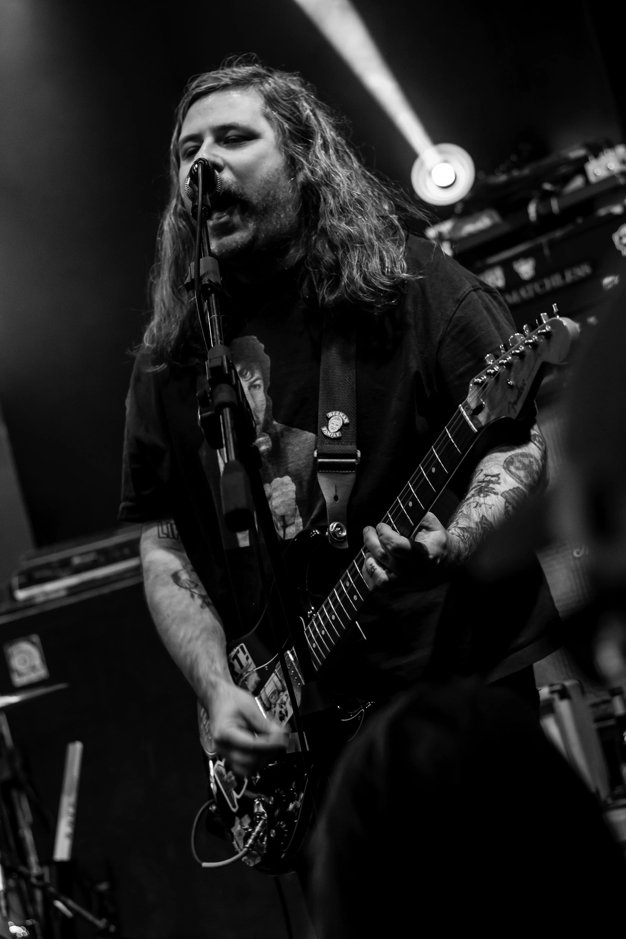 A male musician with long hair and tattoos playing an electric guitar and singing into a microphone on stage in a dimly lit setting.