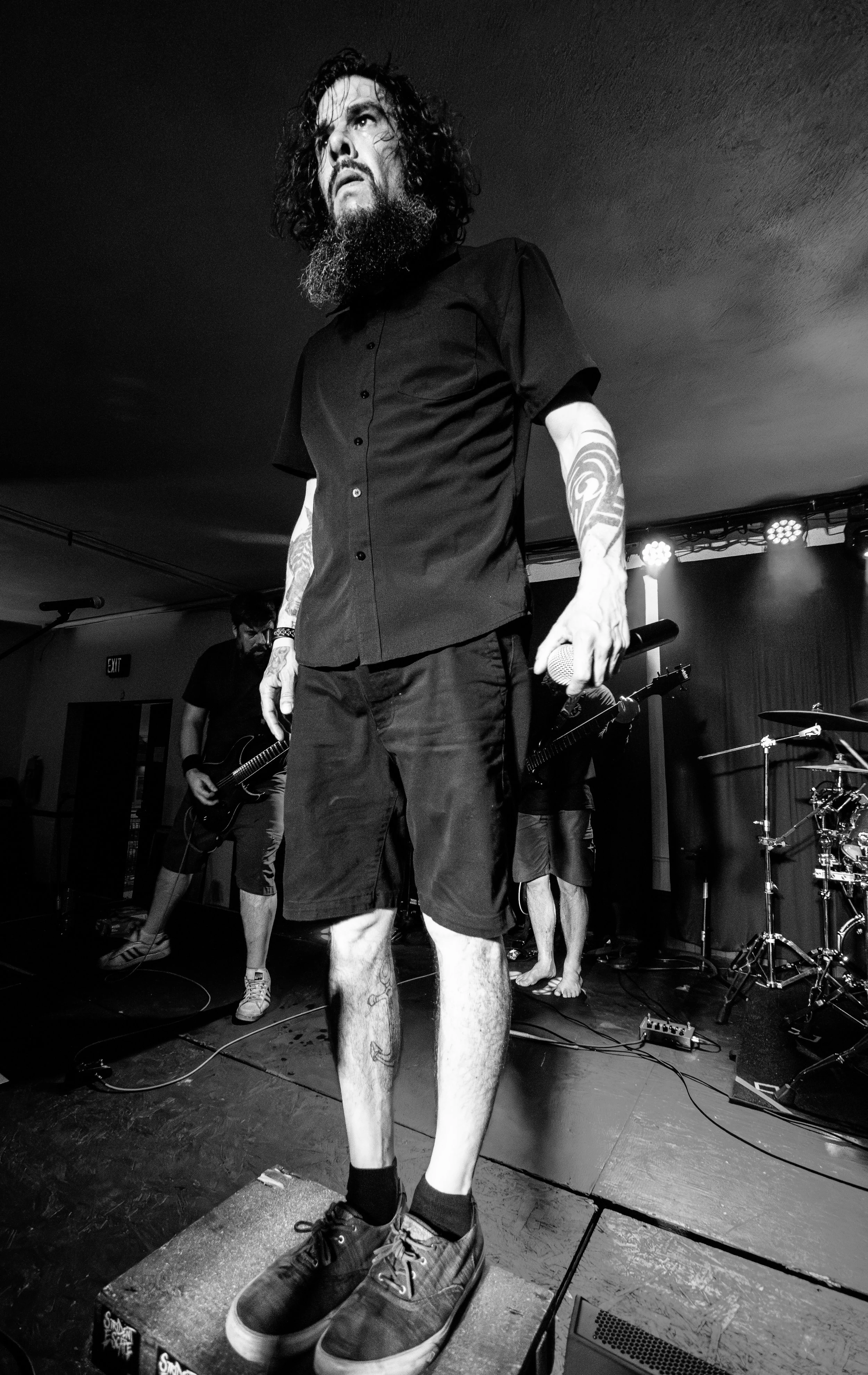 Black and white photo of a man with a beard and tattoos standing on a stage holding a microphone, with a band playing instruments in the background.