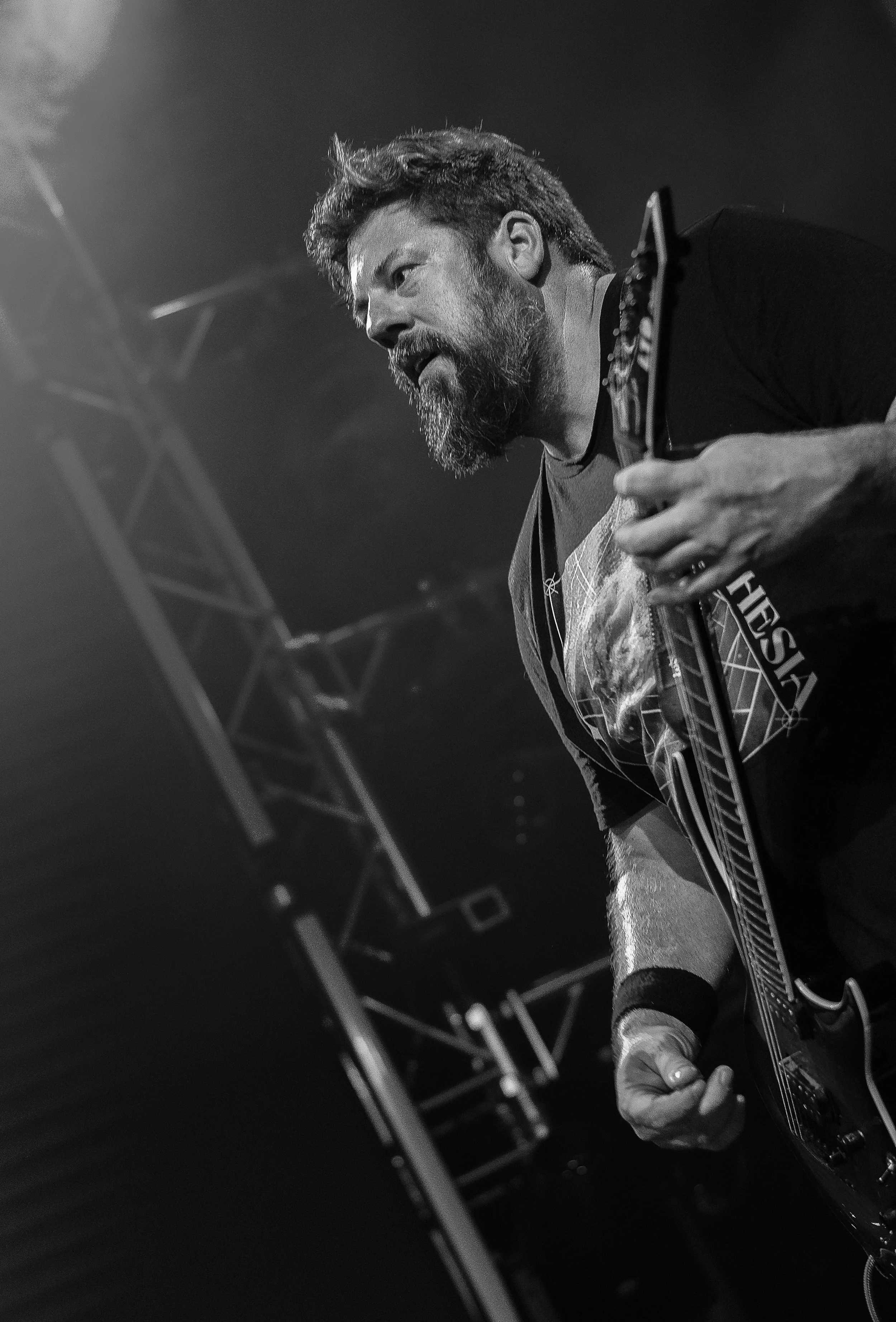 Black and white close-up of a male guitarist playing an electric guitar on stage, with stage lighting and metal scaffolding in the background.