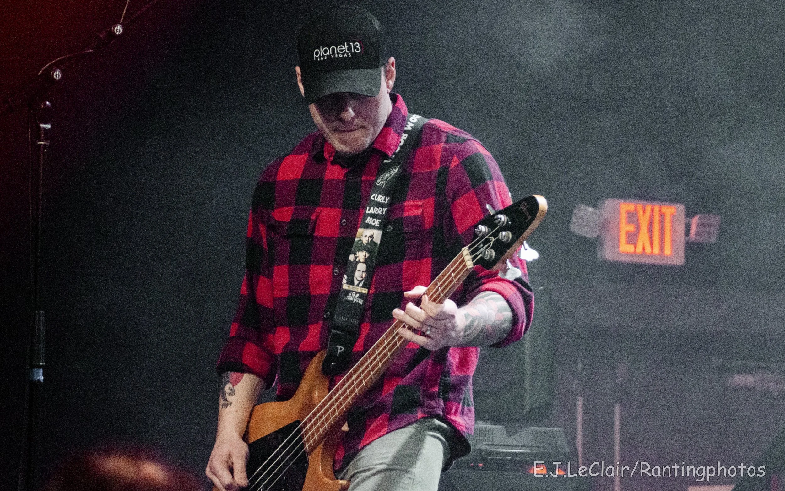 A person playing a guitar on stage, wearing a red and black plaid shirt, a black cap with 'planet13 Las Vegas' logo, and a black lanyard with photos. The background is dark with an illuminated exit sign.