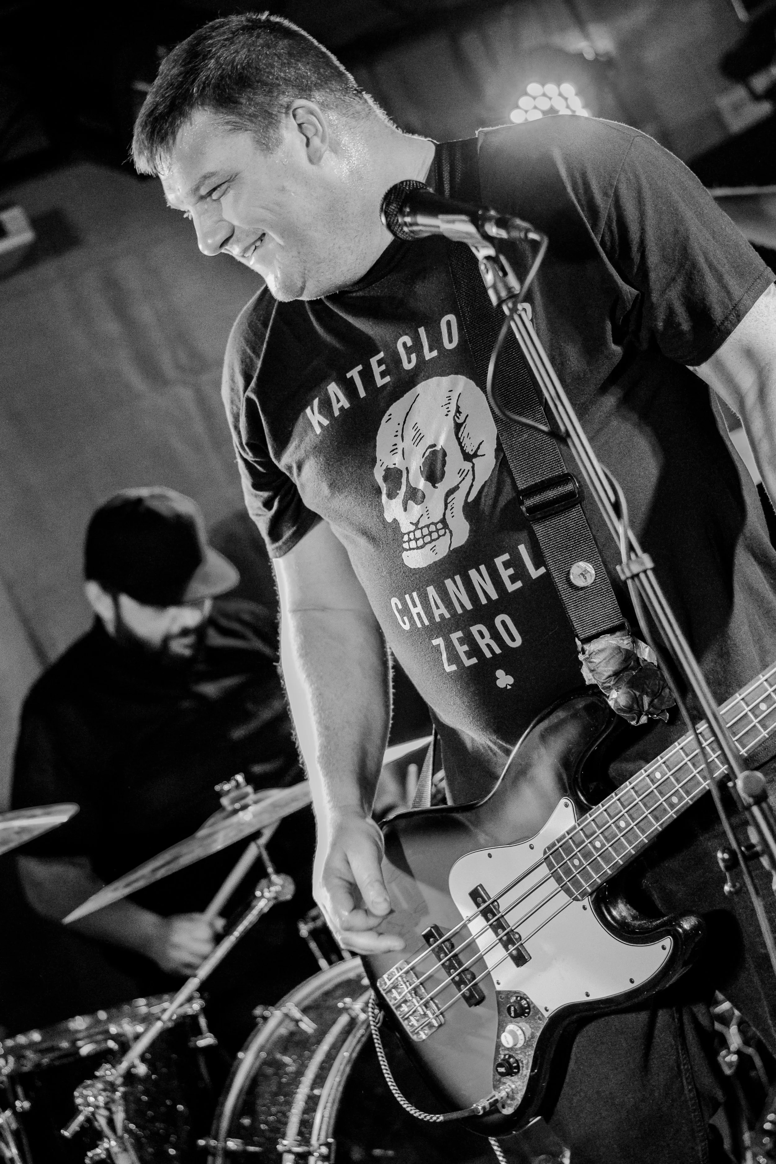 A man playing an electric guitar on stage, smiling with his body leaning forward. He is wearing a T-shirt with a skull graphic and the words 'KATELO CHANNEL ZERO.' There is a microphone near his mouth, and another person is visible in the background 