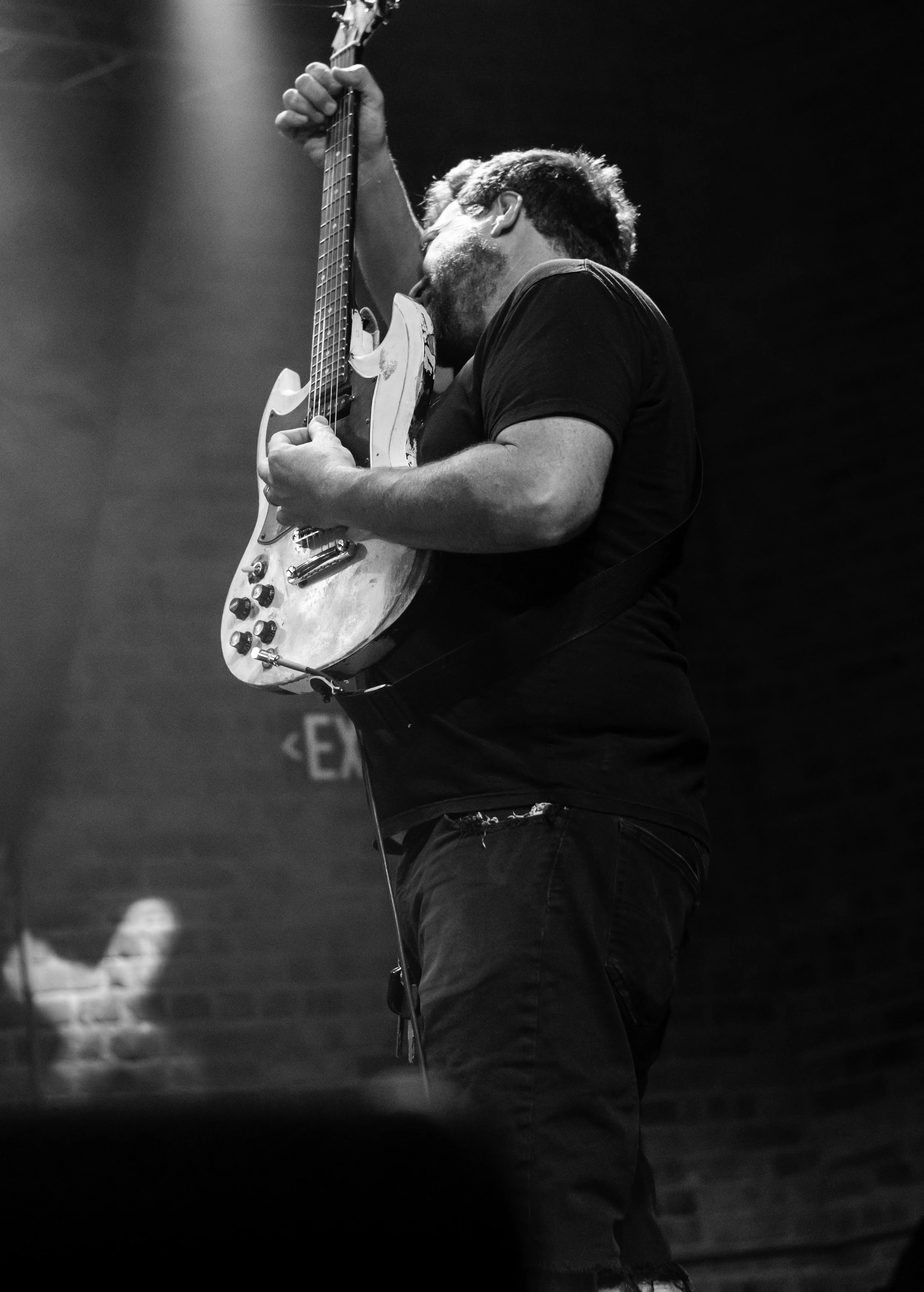 A man playing an electric guitar on stage in a black and white photo, partially obscured by a dark silhouette.