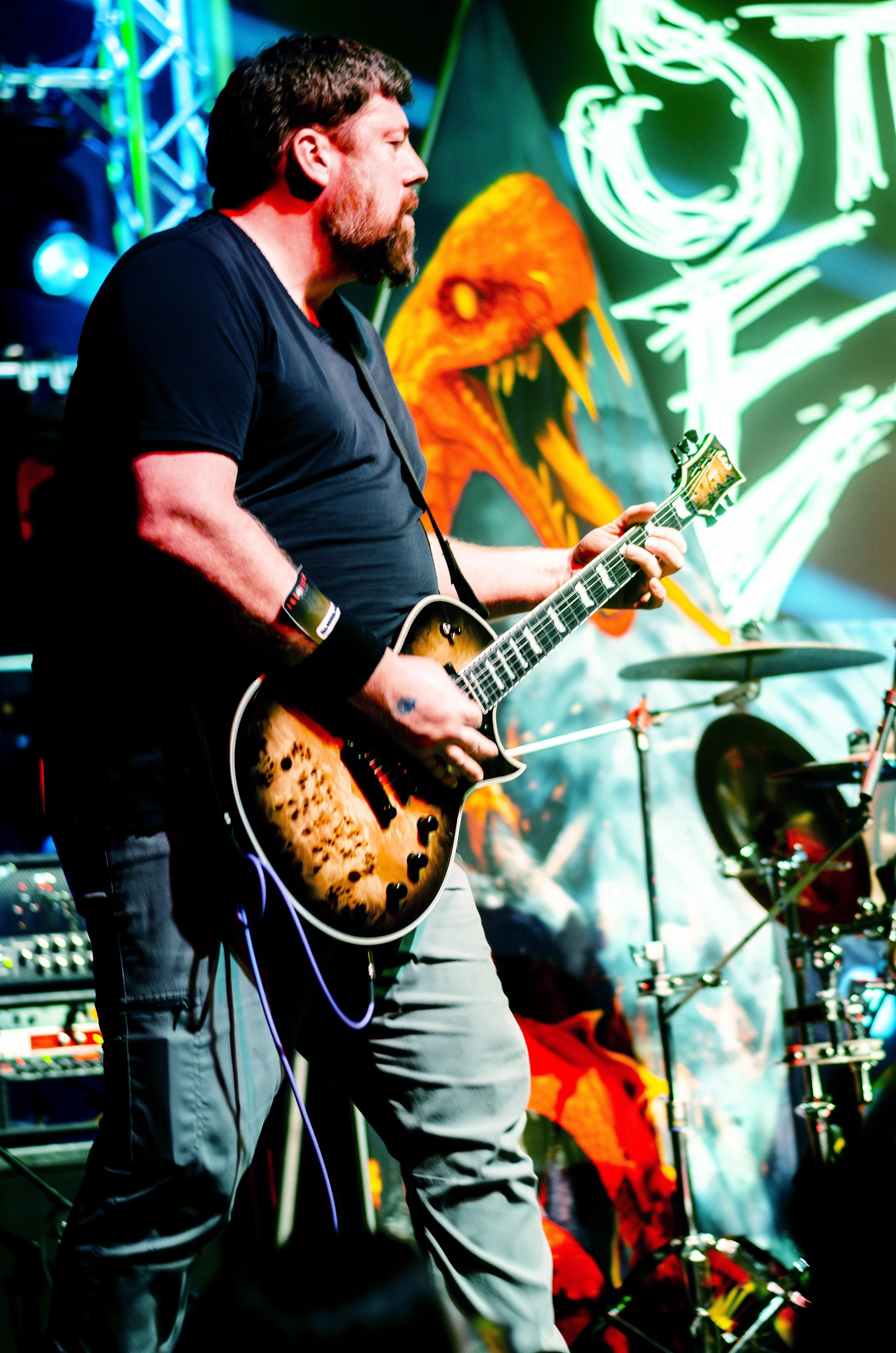A man playing an electric guitar on stage during a live musical performance, with vibrant lighting and a colorful background that includes a large mural or painting.