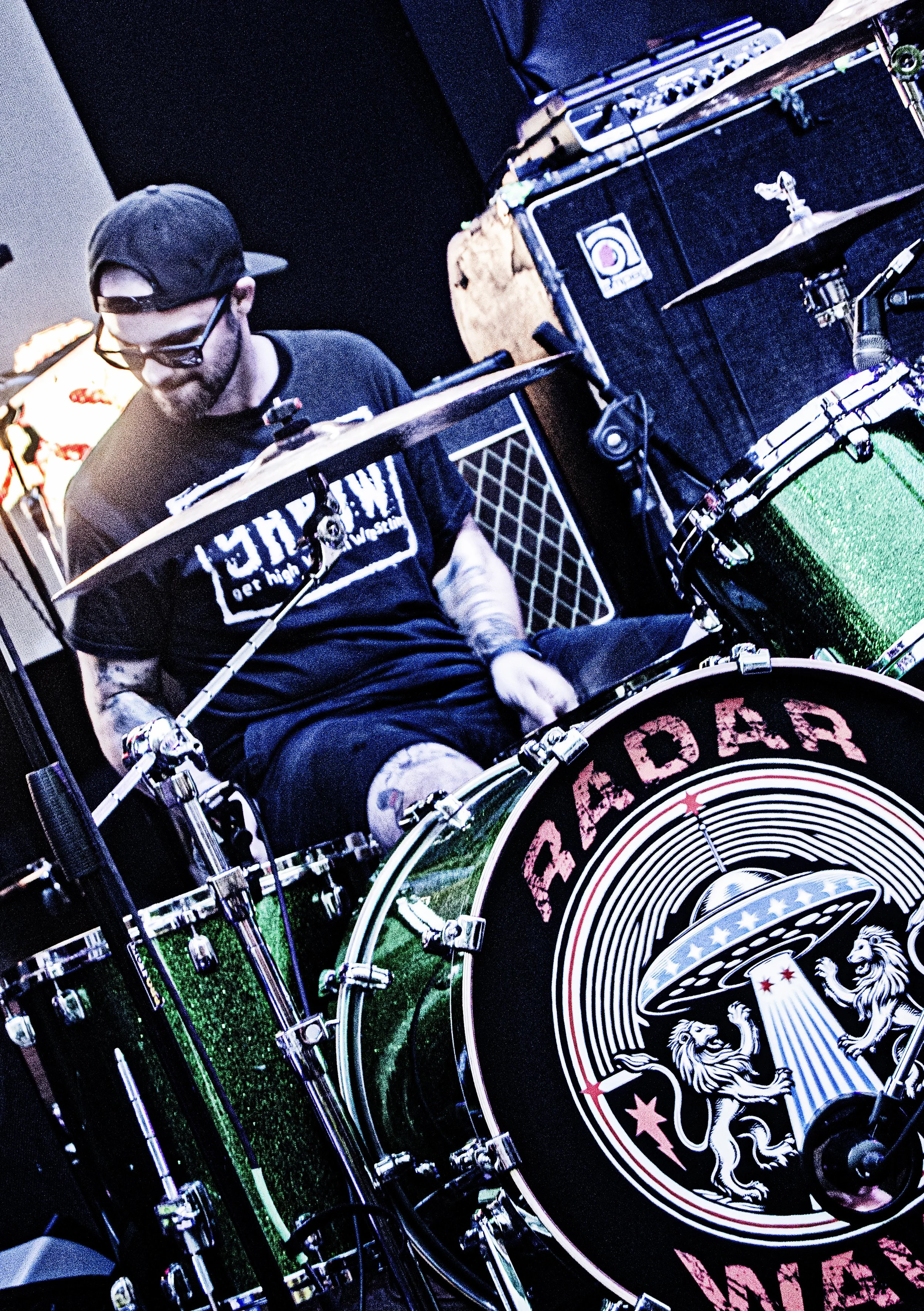 A person with a beard, wearing a black cap and sunglasses, is sitting behind a drum set with a black bass drum that has a logo featuring a lion, stars, and a white circular design. The person is wearing a black t-shirt and has visible tattoos on thei