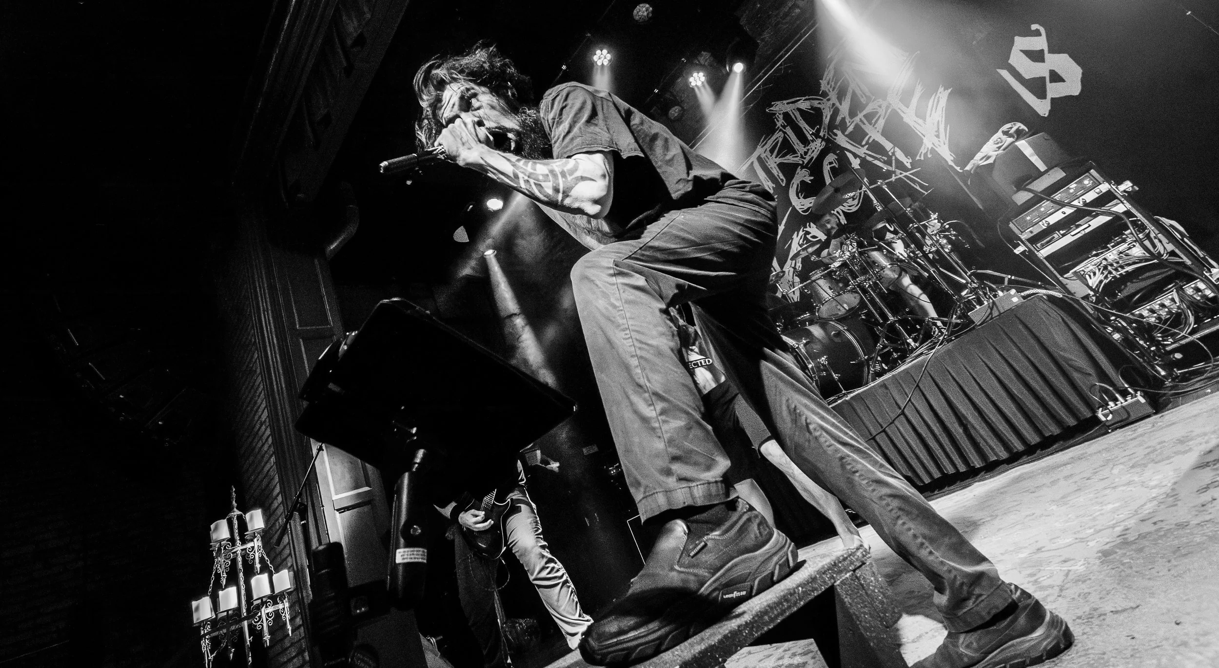 A black and white photo of a musician singing into a microphone on stage with a drummer and guitarist behind him, with stage lights and sound equipment in the background.