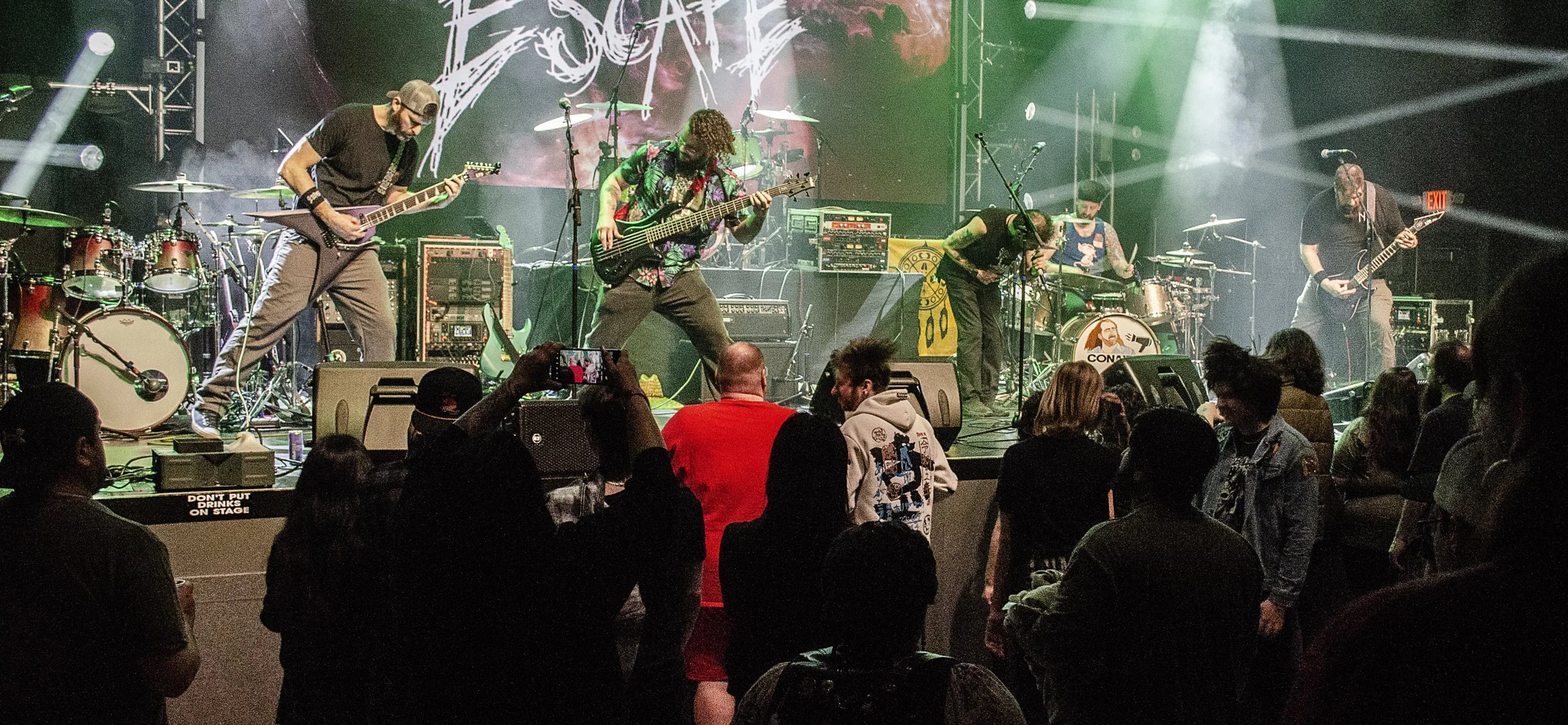 A rock band performing on stage with colorful lighting, featuring five musicians playing guitars and drums, in front of an audience.