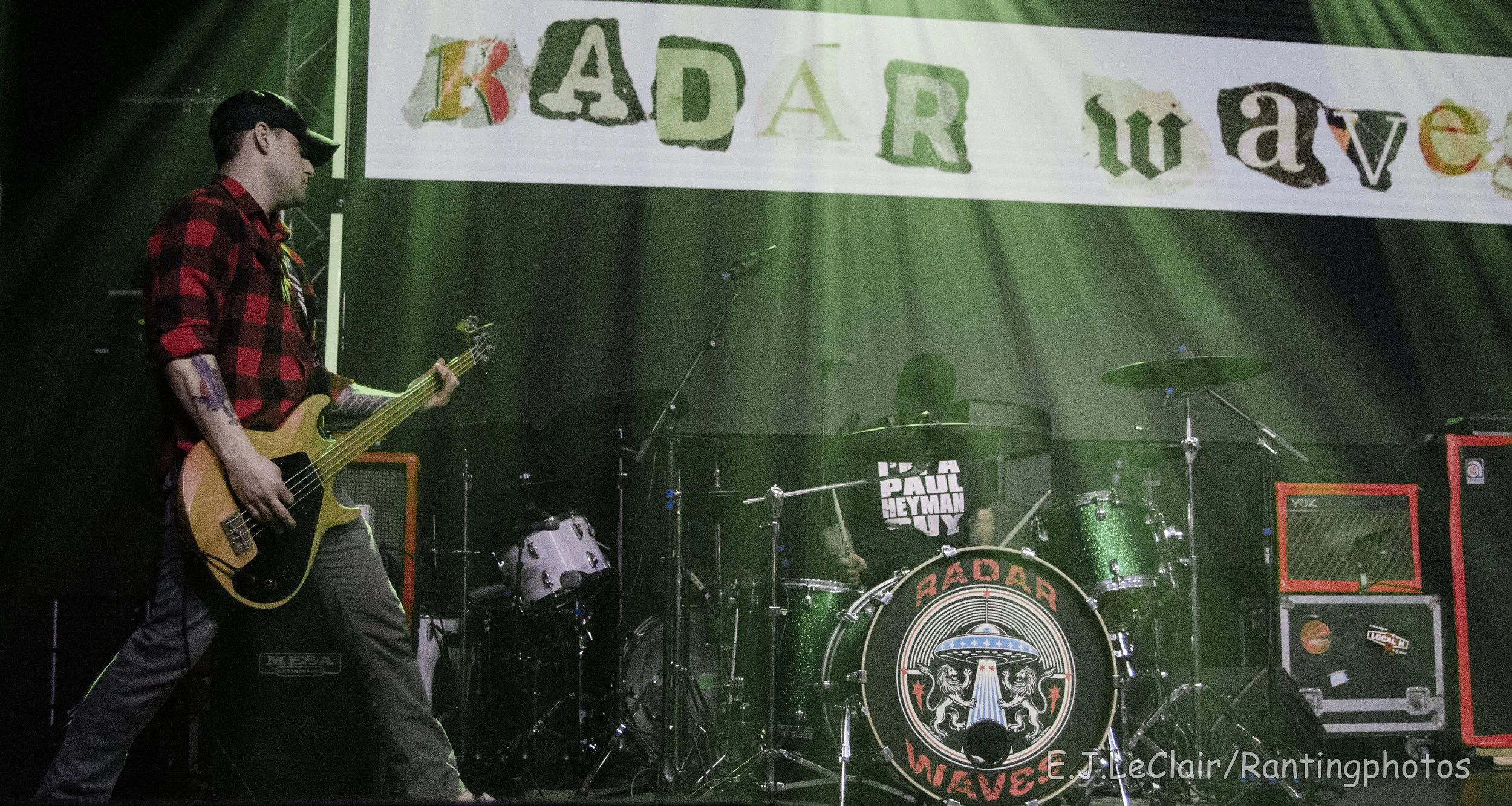 A musician in a red and black plaid shirt and cap playing an electric bass guitar on stage with drum set and amplifiers around, and a large banner that reads 'RADAR' with colorful letters hanging above.