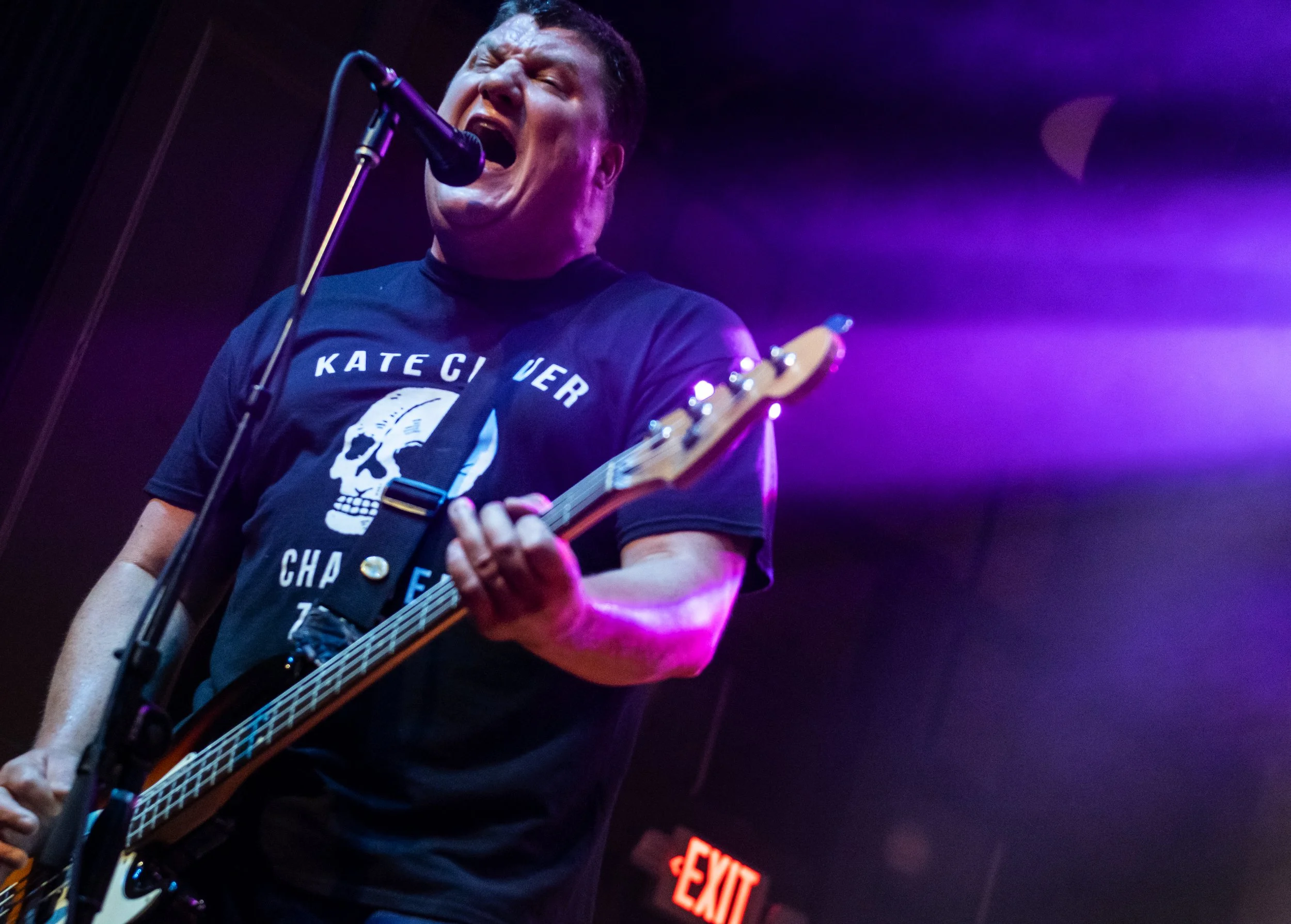 A person singing passionately into a microphone while playing an electric guitar on stage with purple and blue lighting, wearing a black T-shirt with a skull graphic and the person is in front of a dark background with an illuminated exit sign.
