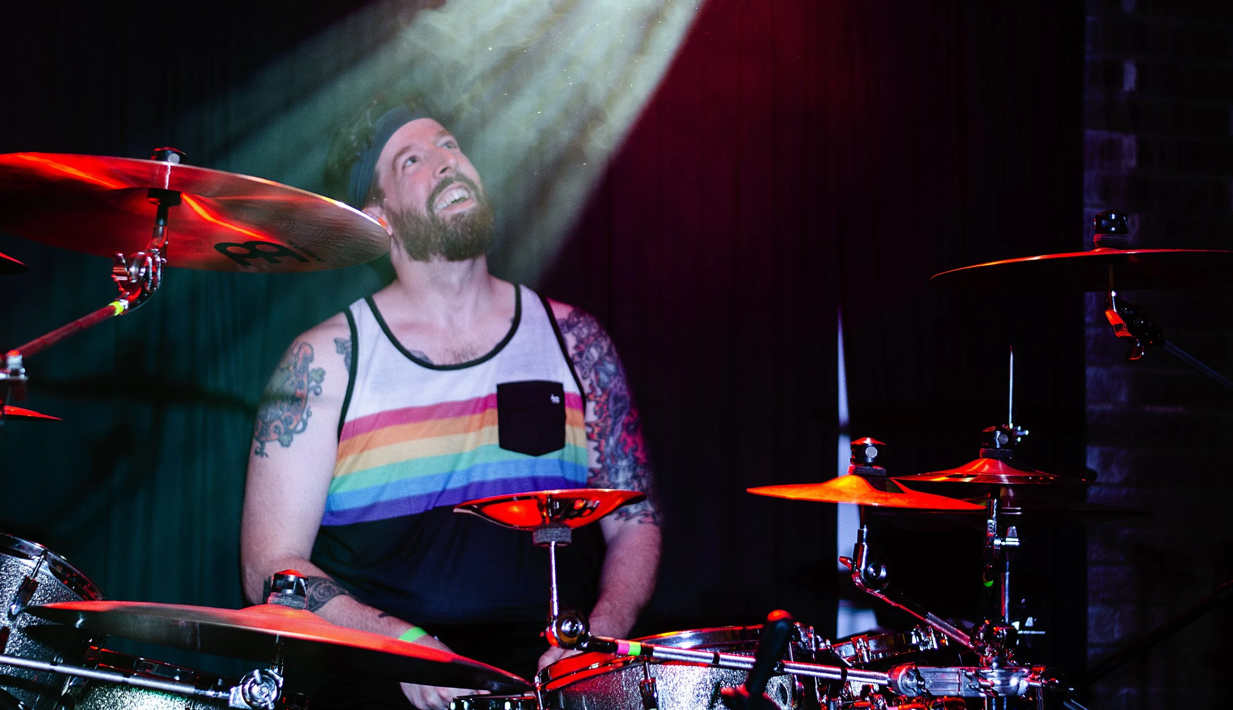A bearded man wearing a rainbow-striped tank top and a black cap playing drums on stage with colorful stage lighting.
