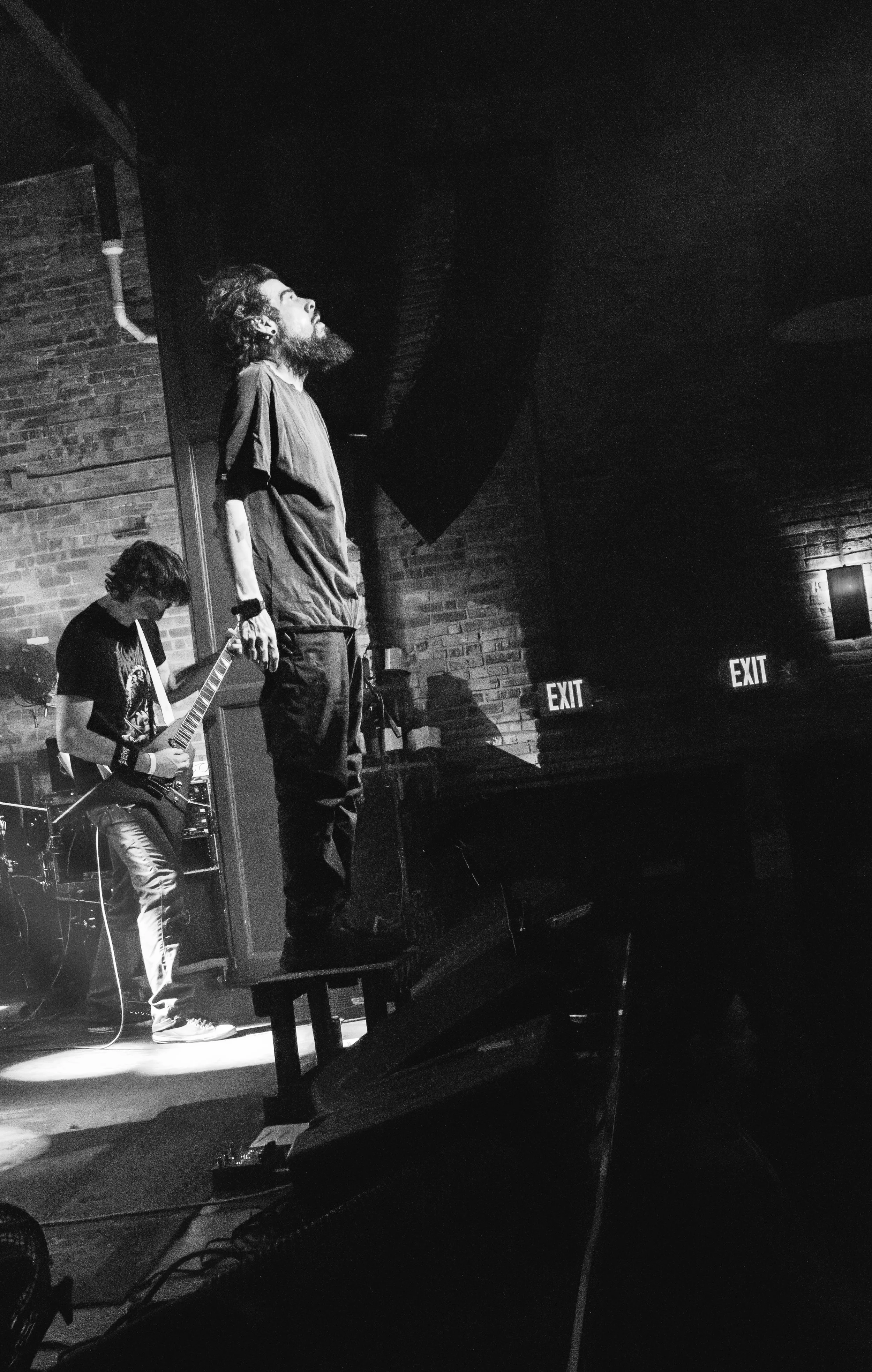 Black and white photo of two musicians performing on a stage with brick wall background; one standing with a beard and long hair, the other playing an electric guitar with long hair, both focused on their performance. Exit signs are visible in the ba