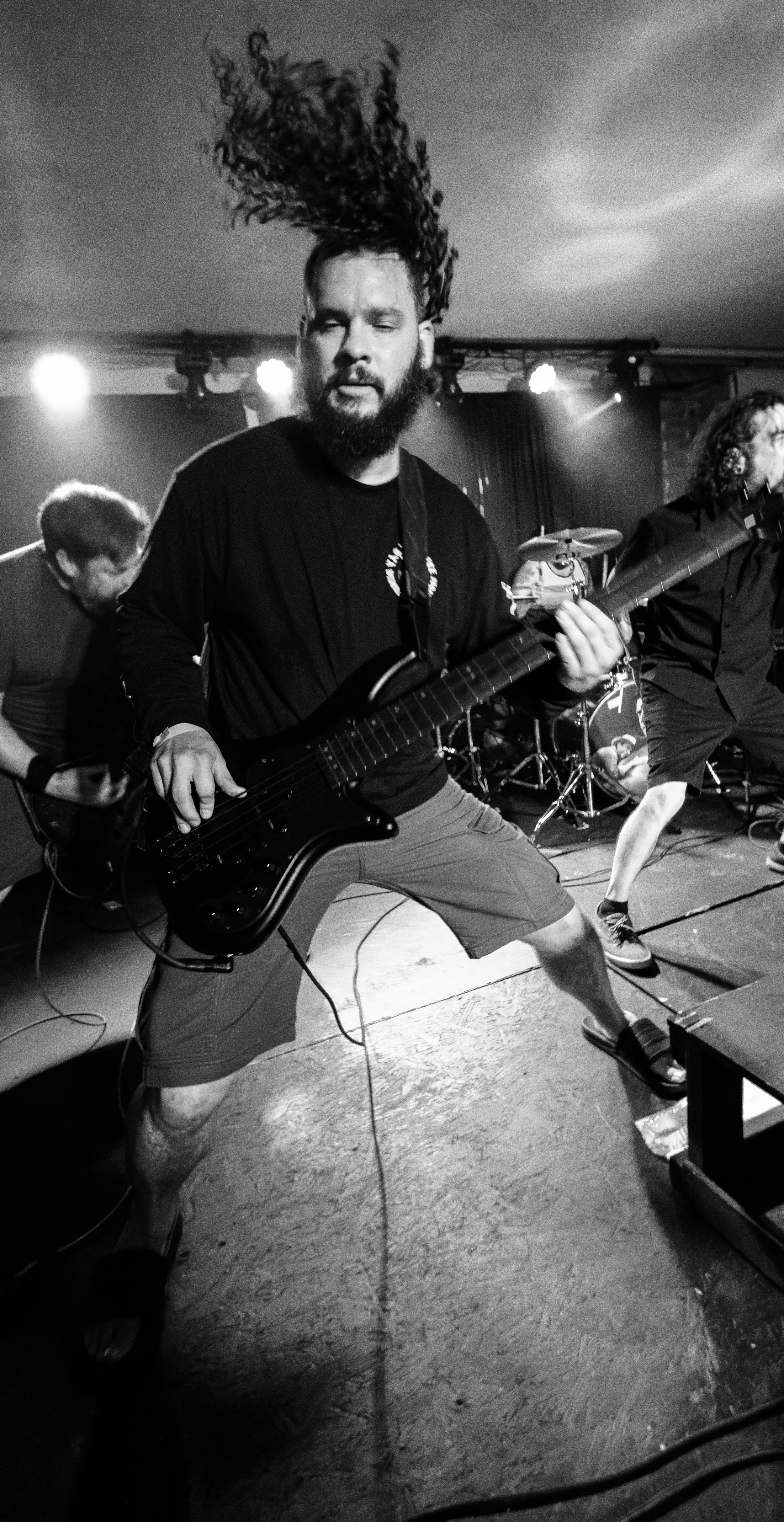 A black and white photo of a band performing on stage, featuring a guitarist with long hair in mid-air, a drummer in the background, and other band members playing instruments.