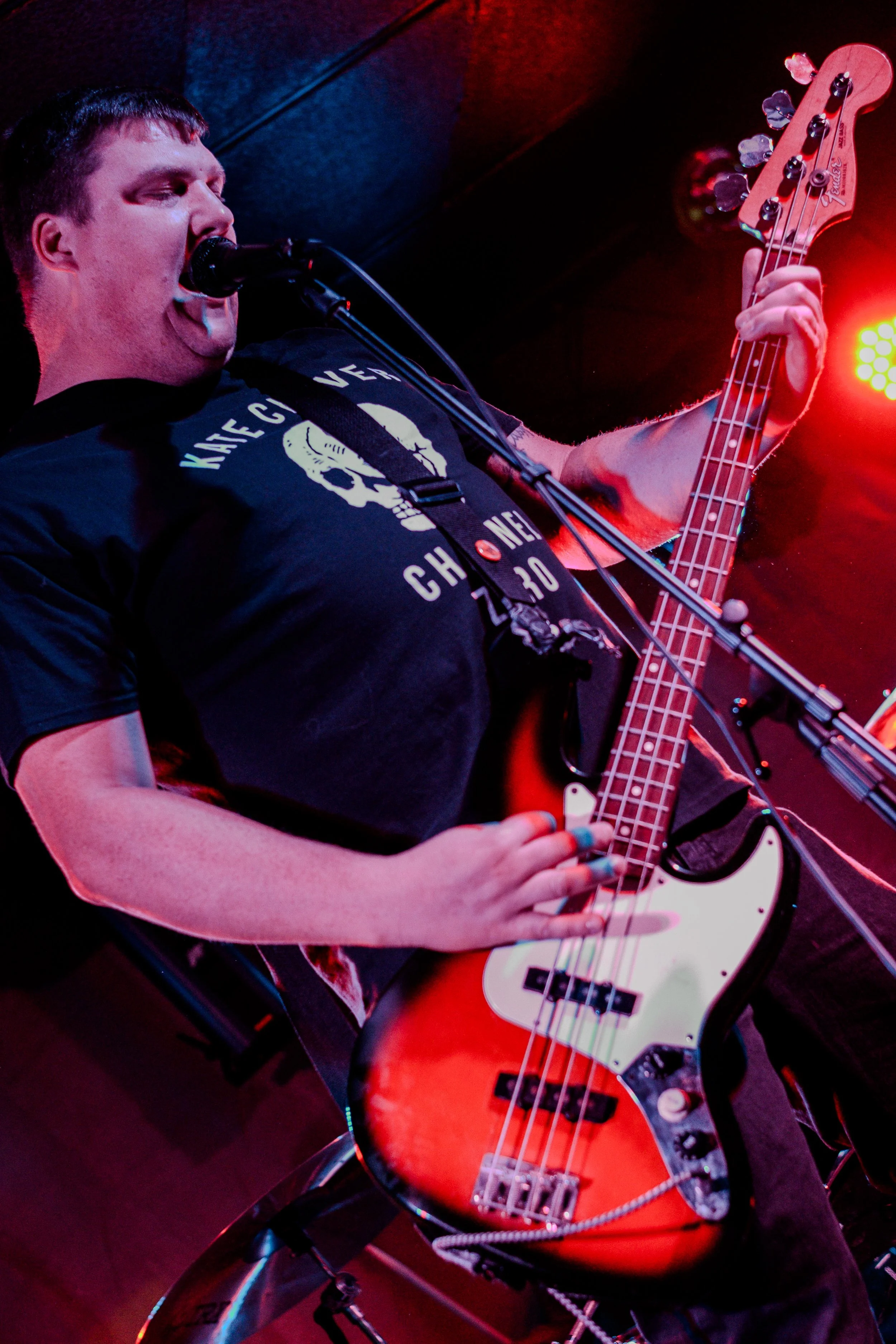 A male musician playing a bass guitar on stage, singing into a microphone, with stage lights creating a red glow.