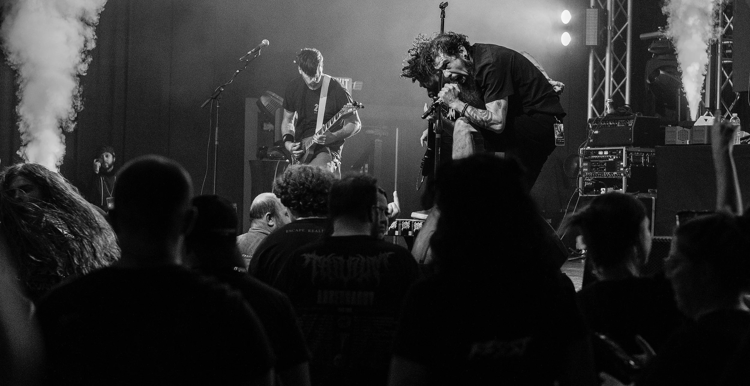Black and white photo of a live music performance with a singer passionately singing into a microphone and a guitarist playing guitar on stage. Audience members are visible in the foreground.