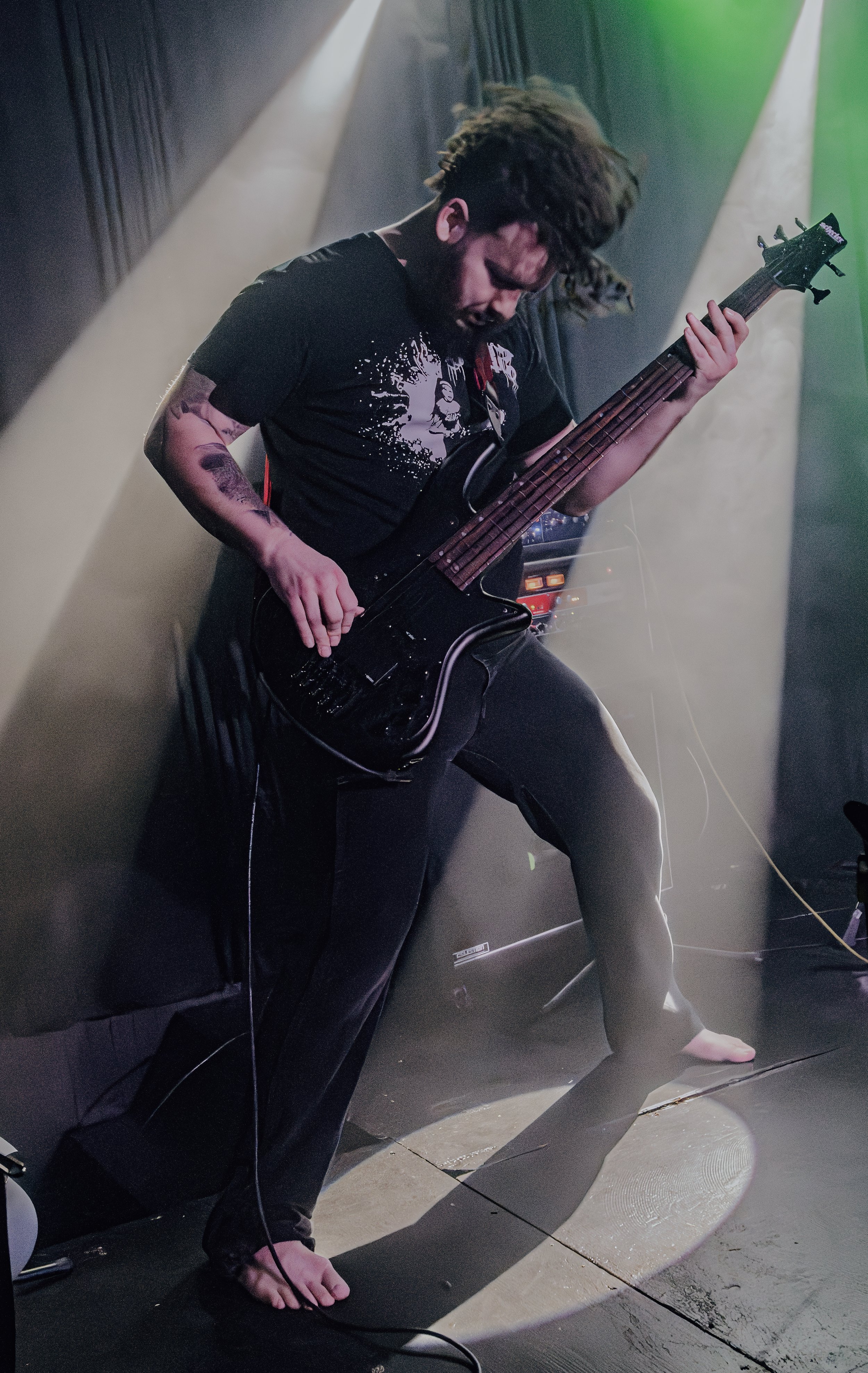 A musician with long hair playing a bass guitar on stage, barefoot, with stage lights and sound equipment visible in the background.
