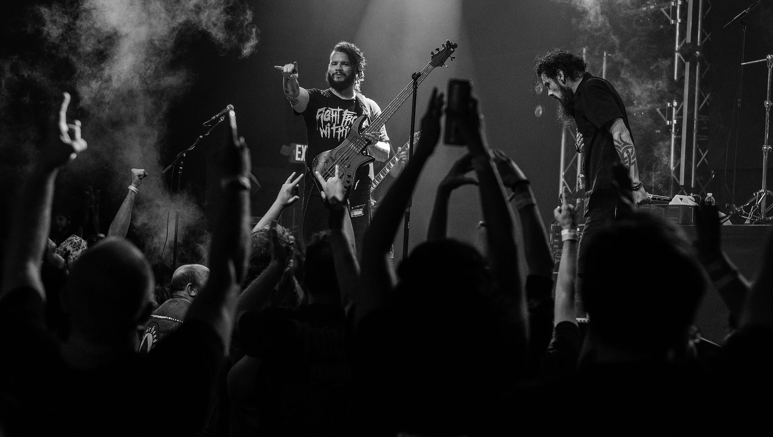 Black and white photo of a live music concert with a band performing on stage, crowd with raised hands, and fog or smoke effects.