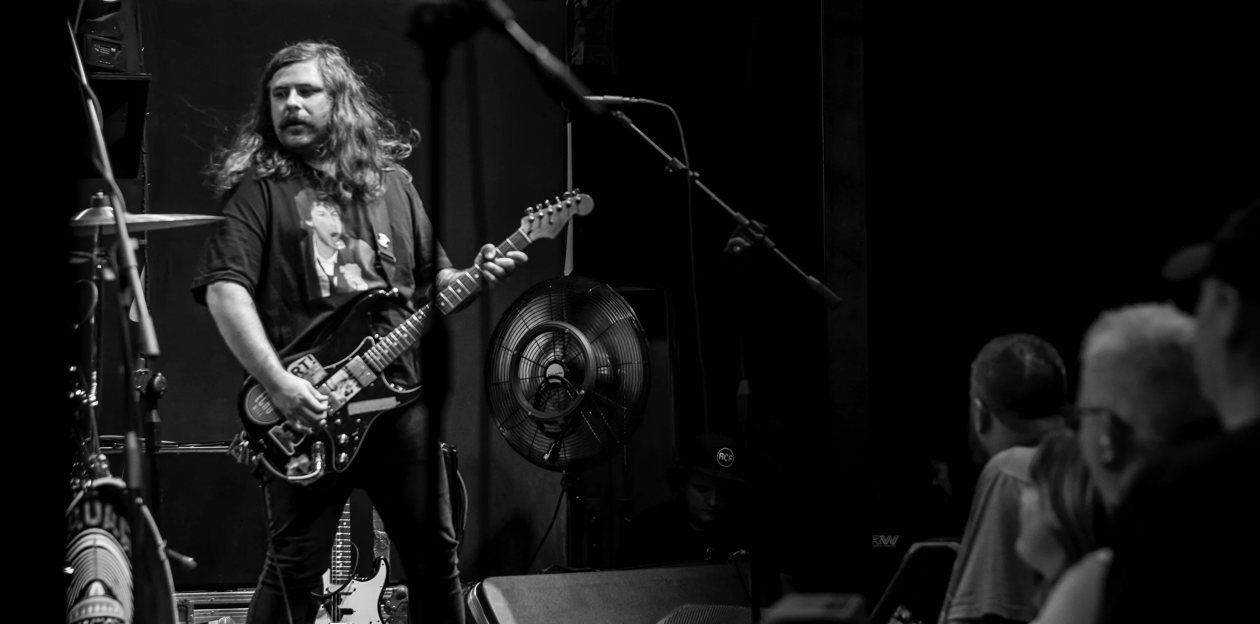 A musician with long hair playing an electric guitar on stage in front of an audience, with a fan and microphone visible