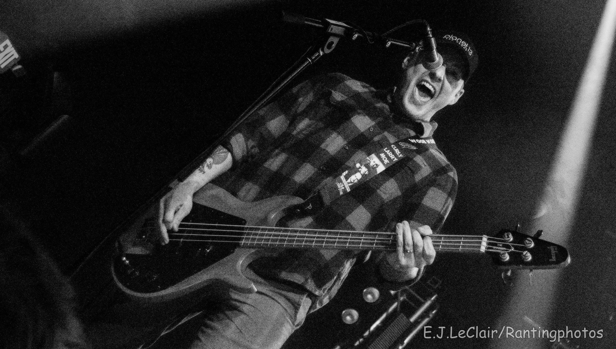 A person wearing a cap and glasses, singing and playing an electric guitar in a dimly lit setting.