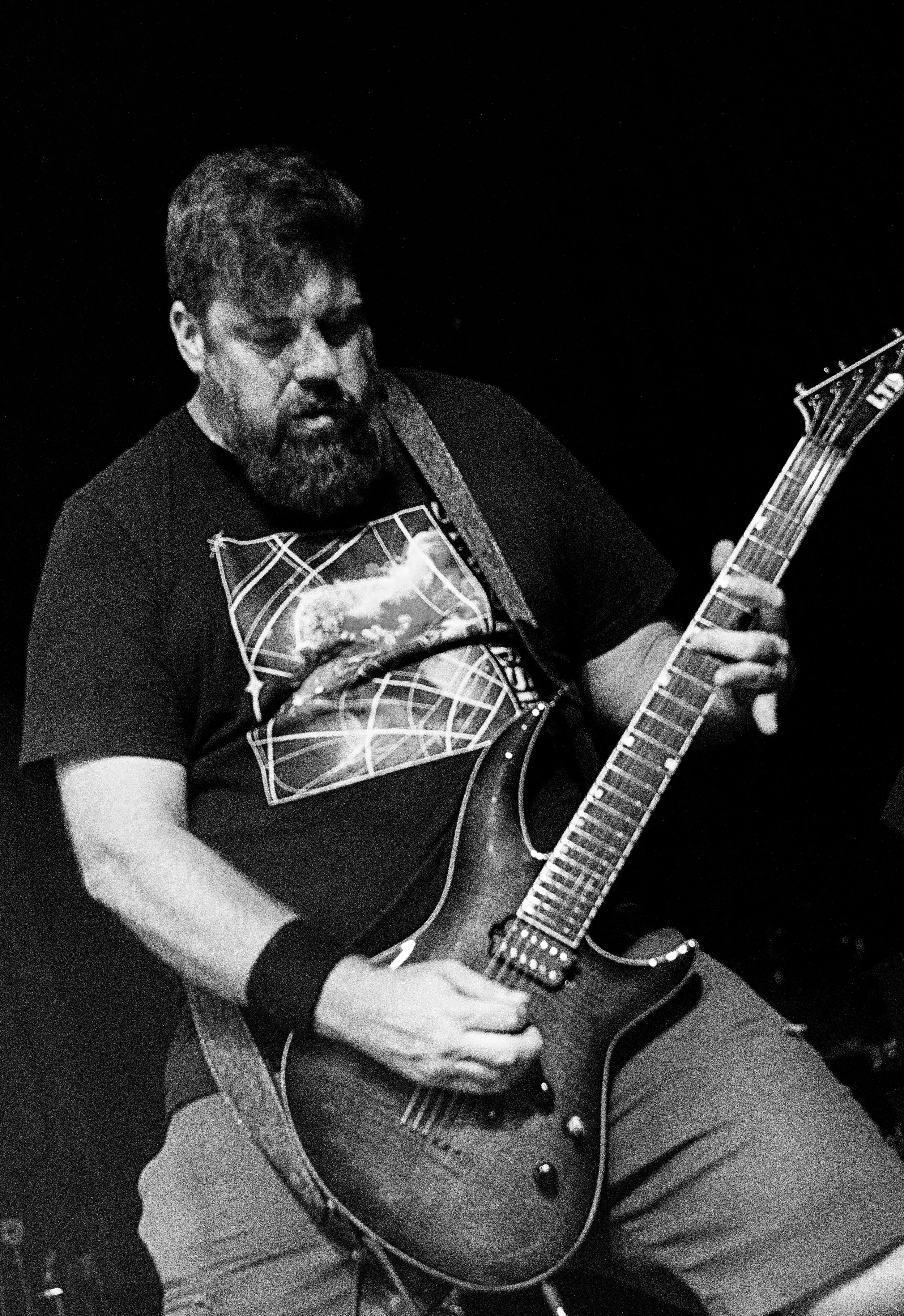 A man with a beard playing an acoustic guitar. He is wearing a T-shirt with a graphic design and appears to be focused on his playing.