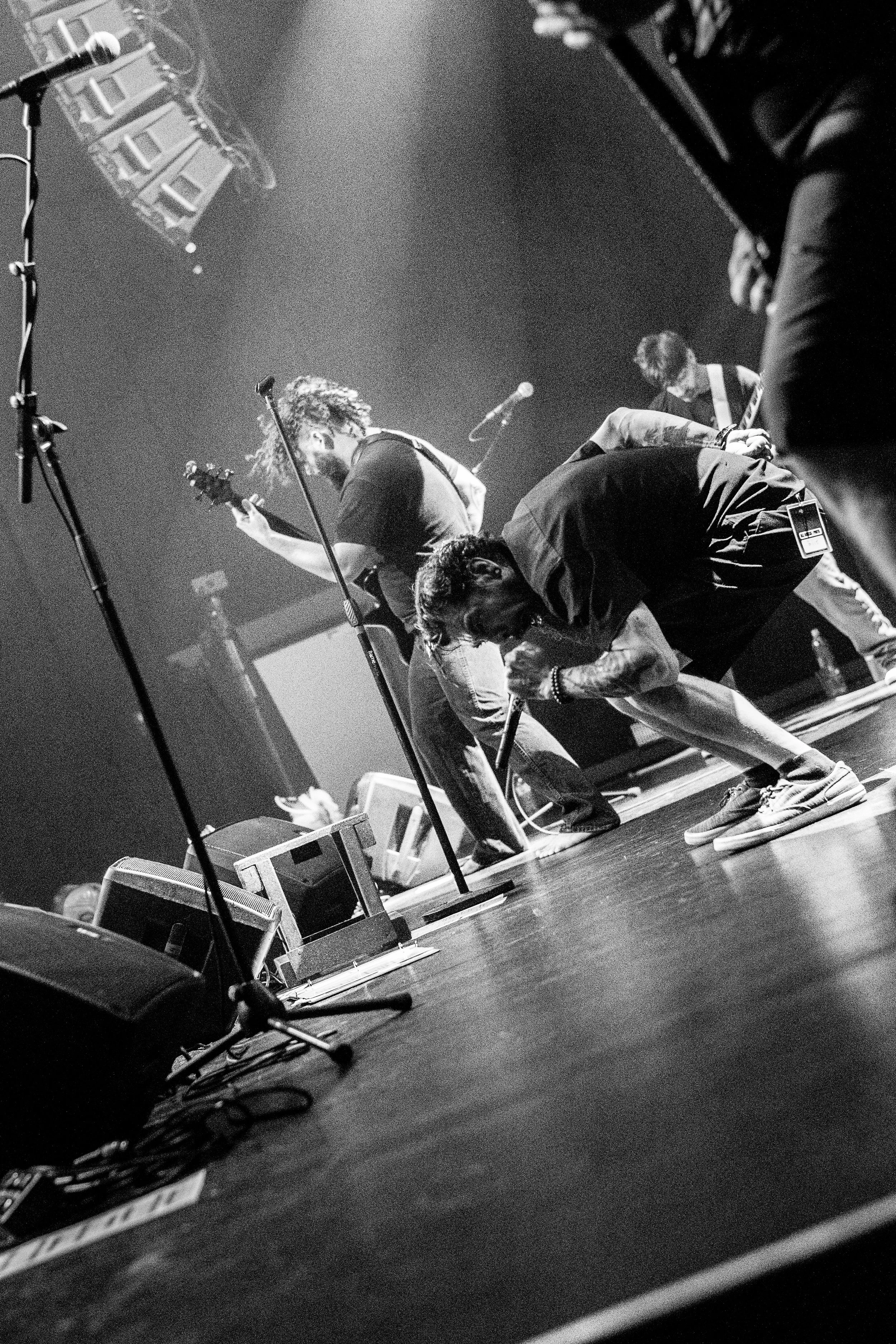 Black and white photo of musicians performing on stage. One person is playing guitar and singing into a microphone, while others are also related to the performance. Equipment and cables are visible on the stage floor.