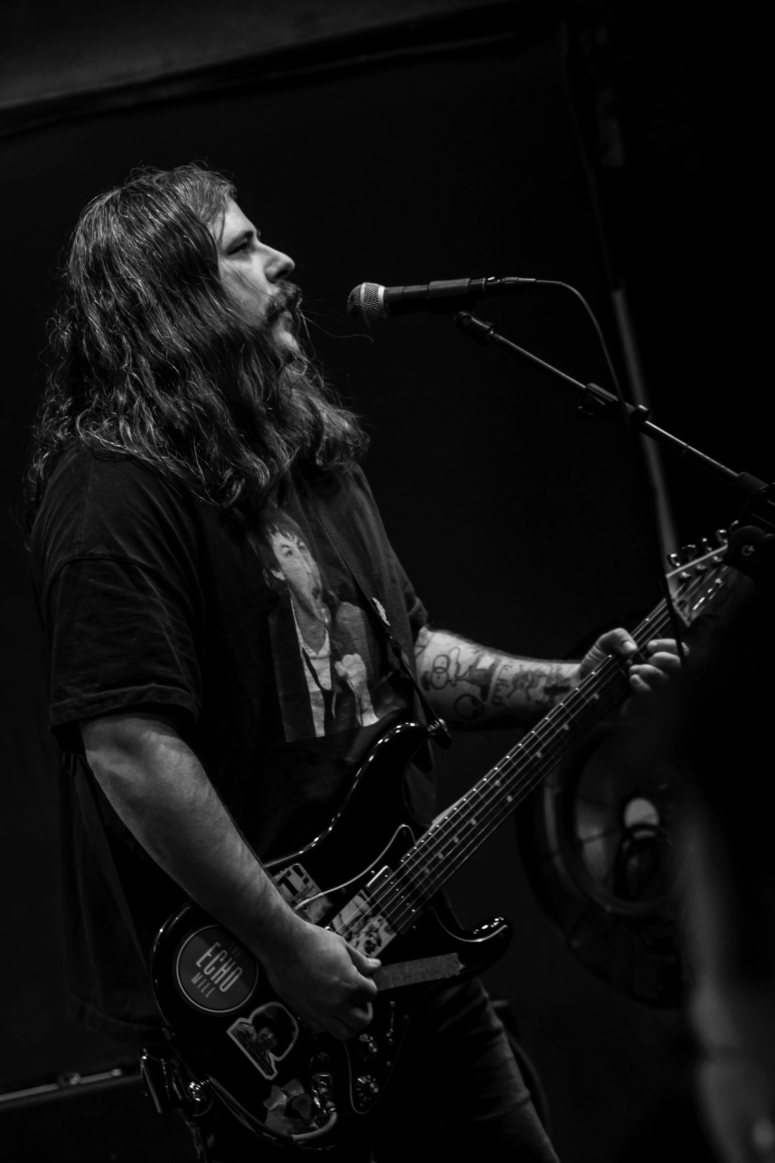 A black-and-white photo of a man with long hair and a beard singing into a microphone while playing an electric guitar.