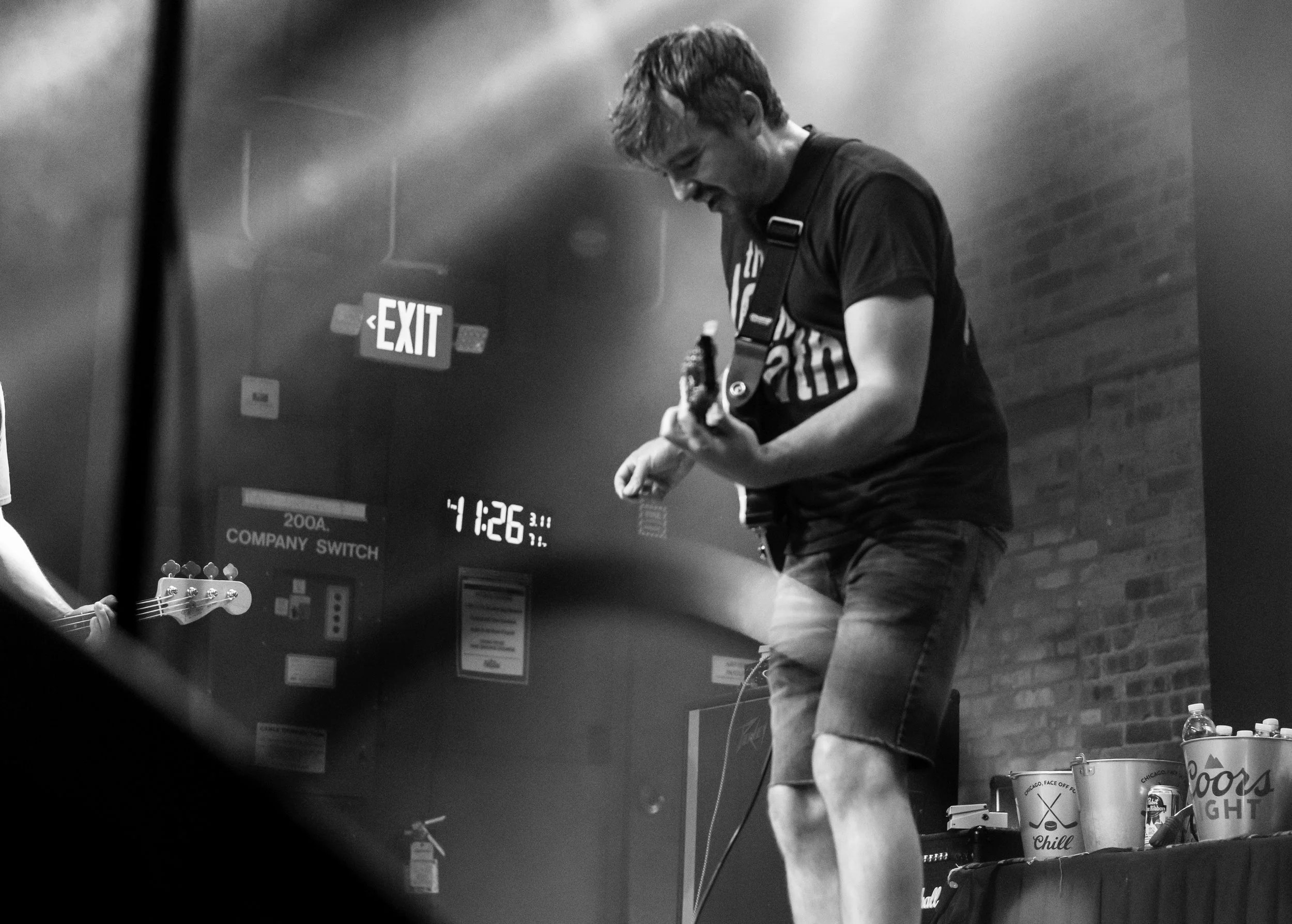 A young man playing an electric guitar on a stage, dressed casually in a t-shirt and shorts, with supplies and a drink nearby, in a black-and-white photograph.