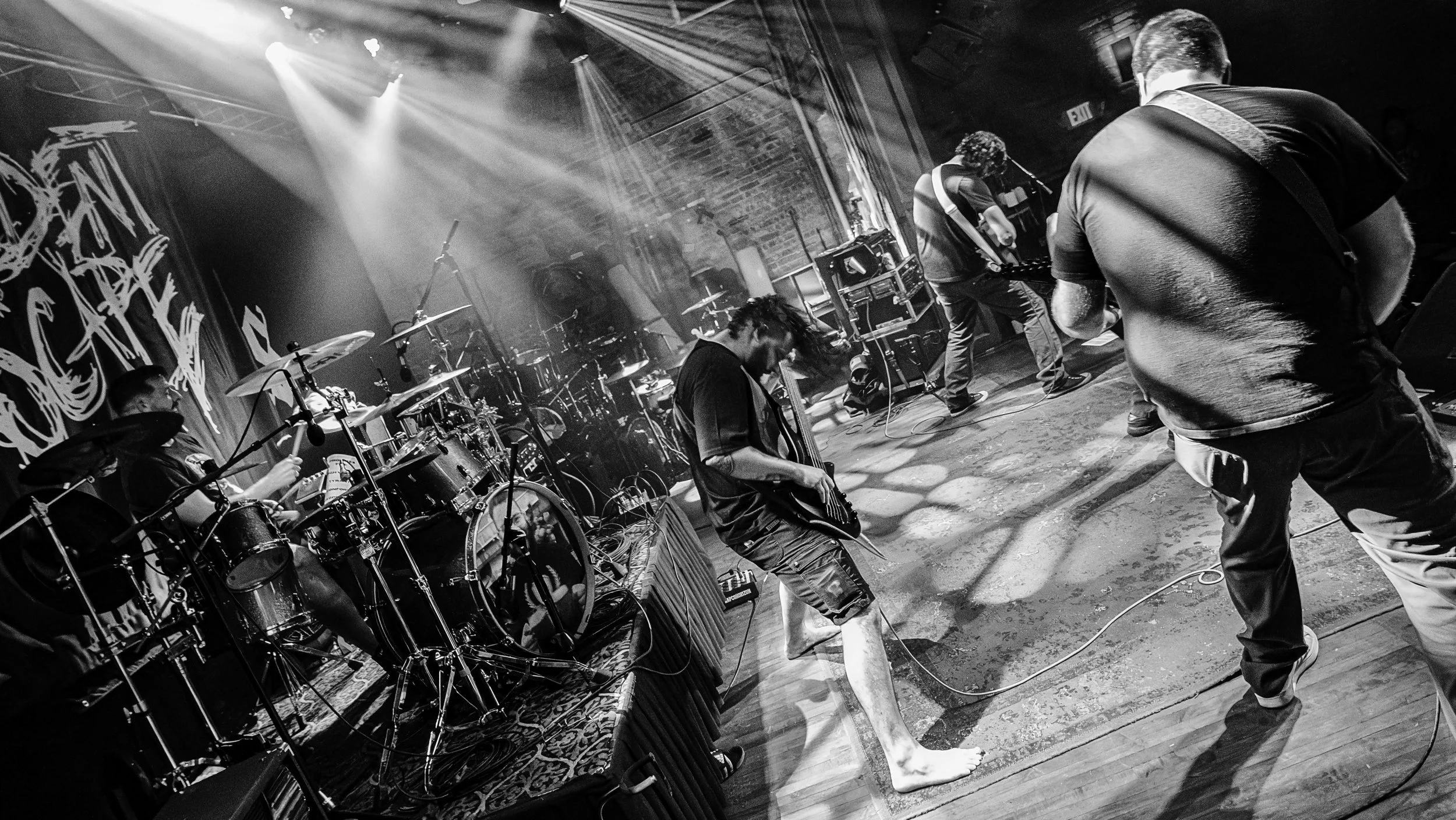 A black and white photo of a band performing on stage. The drummer is to the left, and three guitarists are on the right, with one barefoot. Bright stage lights shine down, illuminating the scene. The background features a brick wall and stage equipm