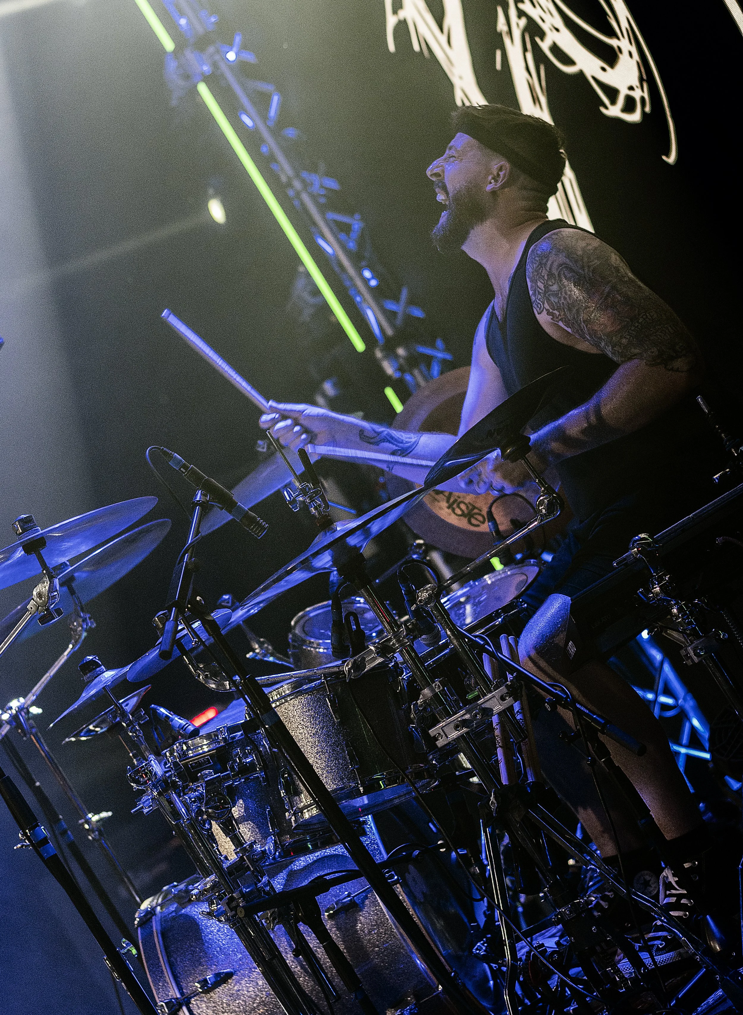 A musician with tattoos on his arm, wearing a black headband and sleeveless shirt, is playing drums on stage with a smile, illuminated by stage lights, and a black backdrop with white graphic designs.