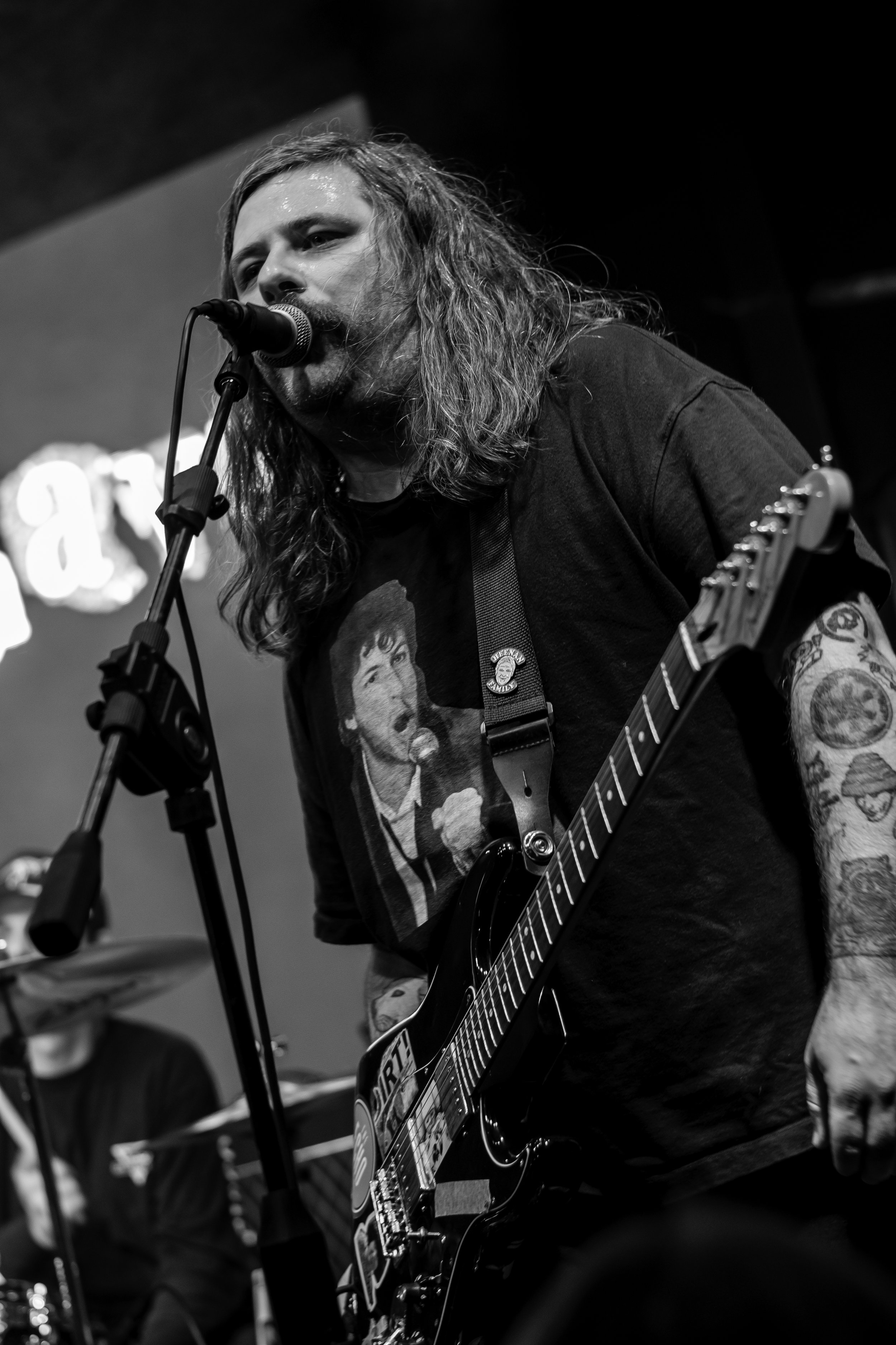 Man with long hair singing into a microphone and playing an electric guitar in a black and white photo.