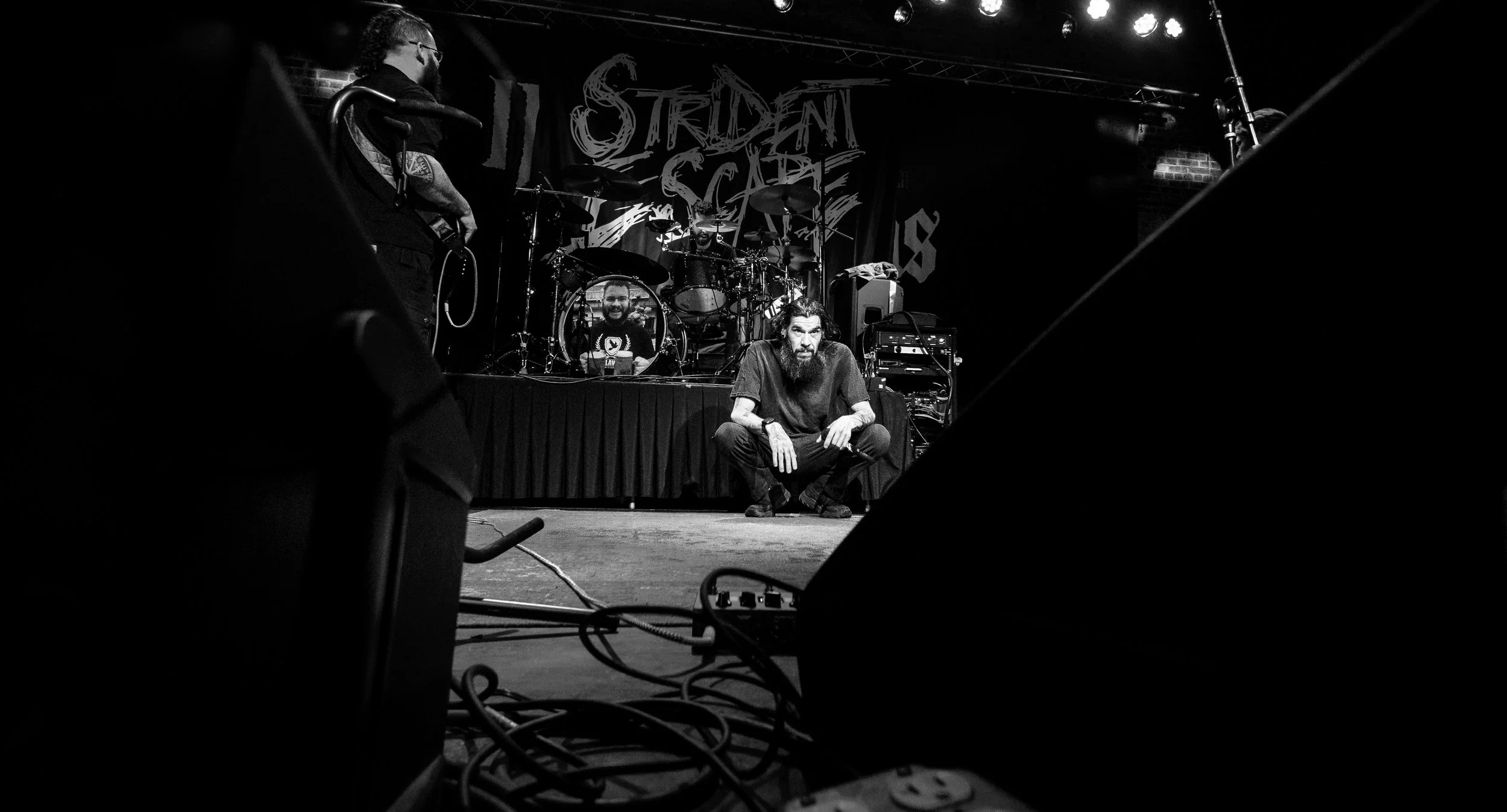 A black and white photo of a band on stage, featuring a male with a beard and long hair crouching in front of the drum kit, a drummer behind him, and a guitarist on the left, with a backdrop displaying "Ordient Scape."