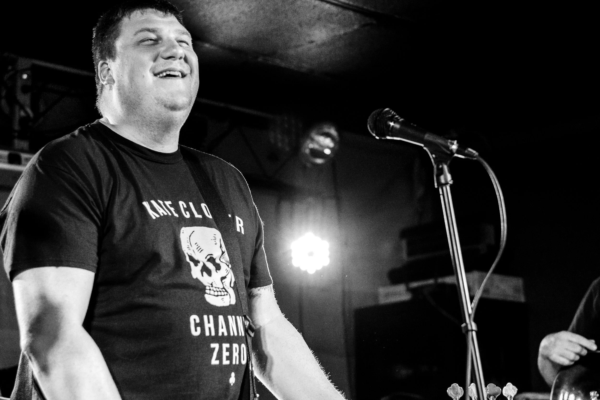 A man singing or speaking into a microphone during a performance, wearing a T-shirt with a skull graphic and text, in a dimly lit setting.