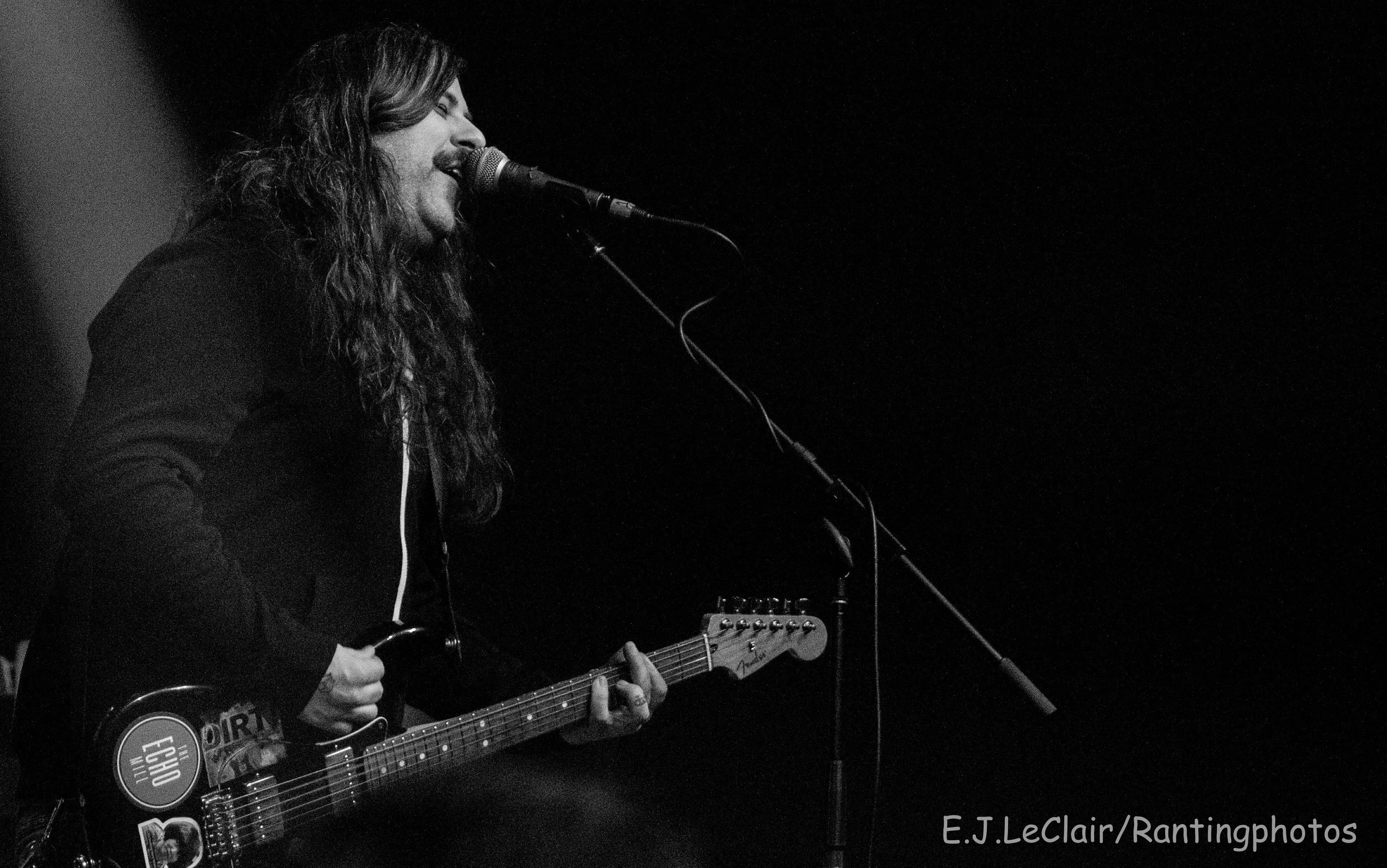 A black and white photo of a man with long hair singing into a microphone while playing an electric guitar on stage.