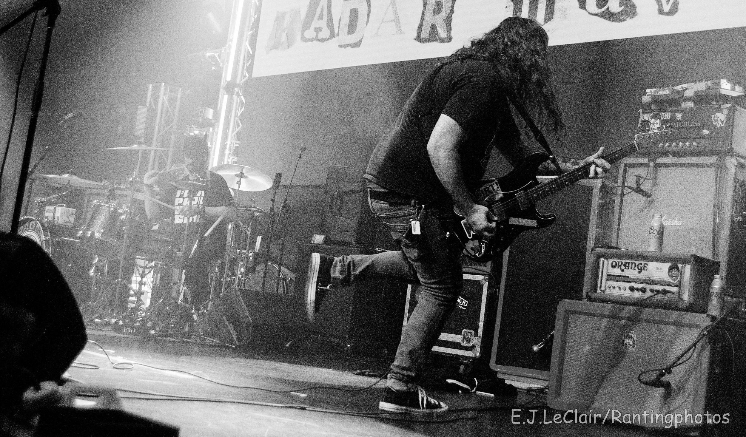 A musician with long hair playing an electric guitar on stage, with a drum set and amplifiers behind him. The scene is in black and white.