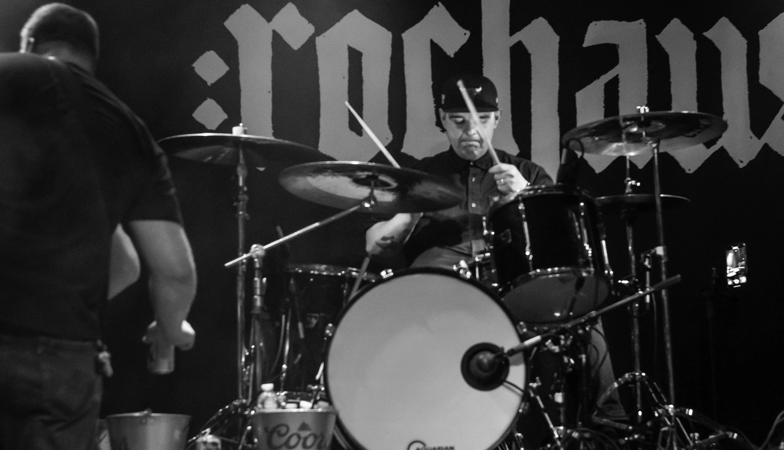 A black-and-white photo of a man playing drums on stage, wearing a helmet and goggles, with a person partially visible in the foreground.