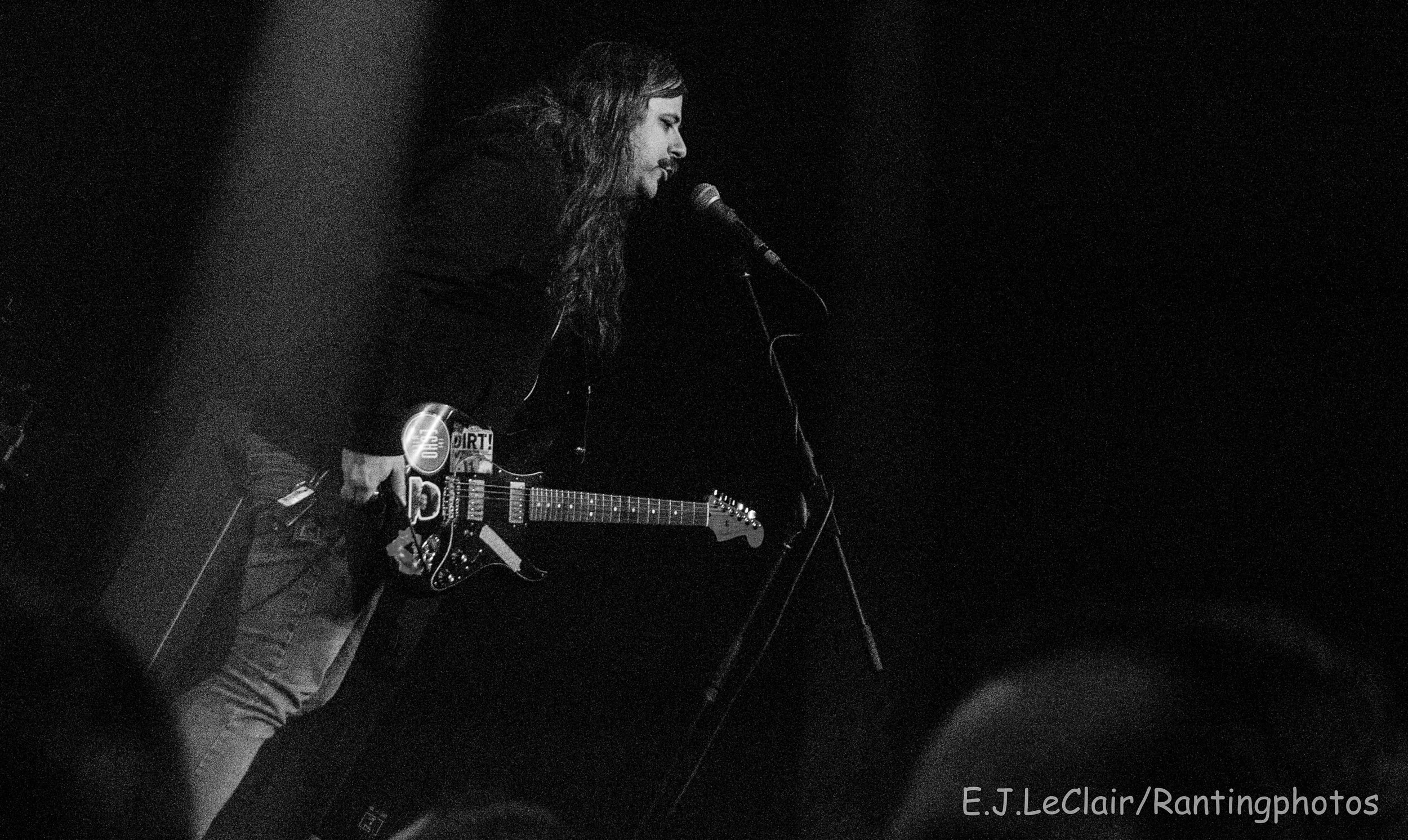A person playing an electric guitar and singing into a microphone on stage, with a dark background.