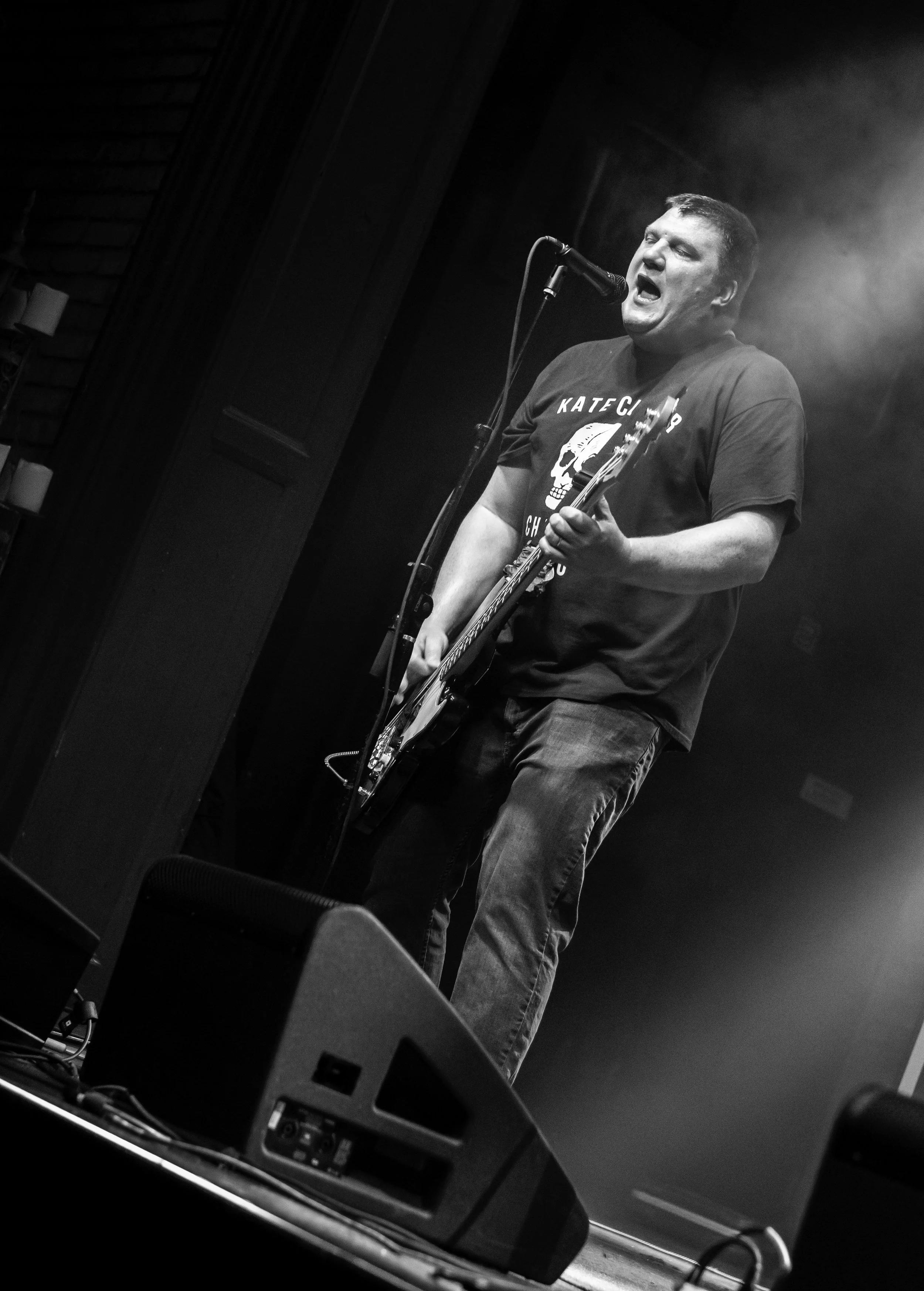 A man singing into a microphone while playing an electric guitar on a stage, with a dark background and smoke effects.