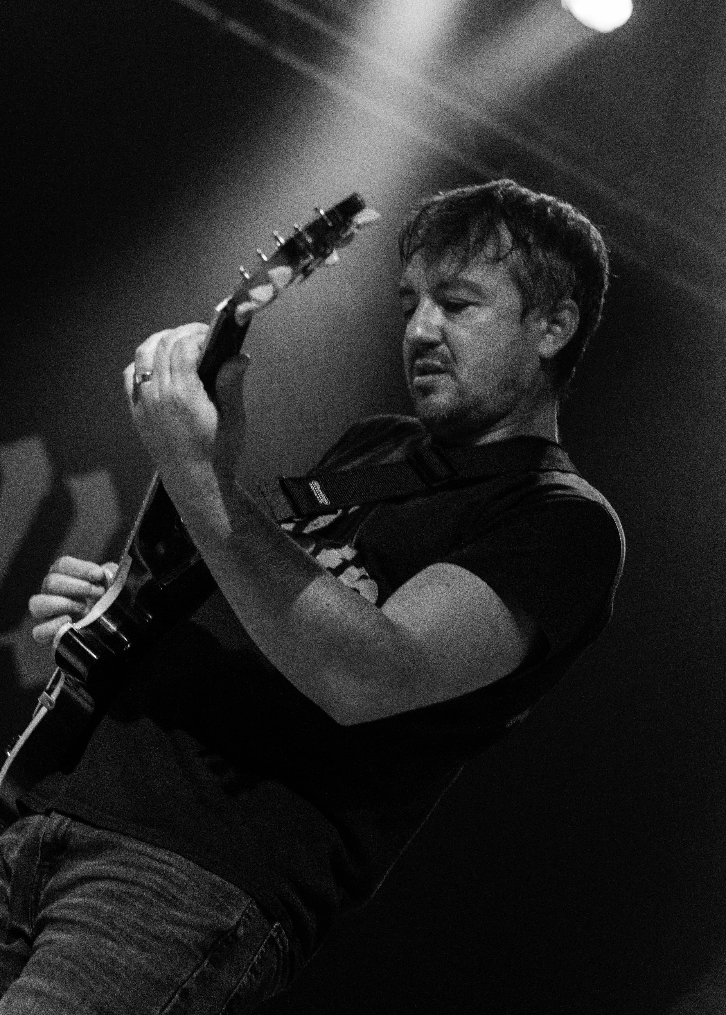 A man playing an electric guitar on stage, with a focused expression, in black and white.
