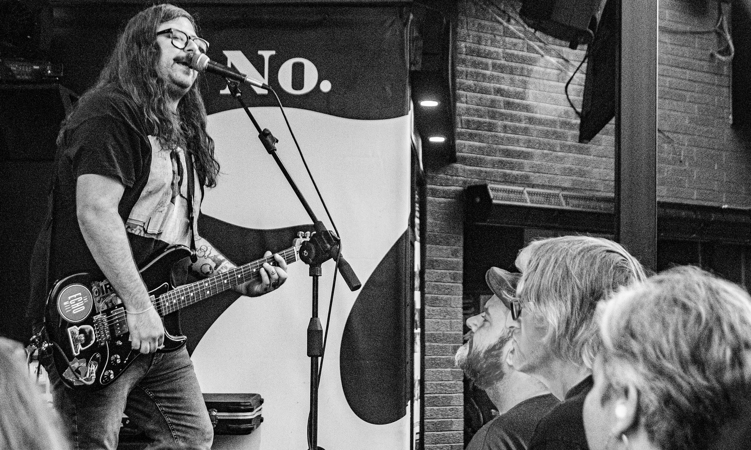 A male musician with long hair and glasses playing an electric guitar and singing into a microphone on stage. The background has a sign with the word "NO." and a brick wall. Audience members are visible in the foreground.