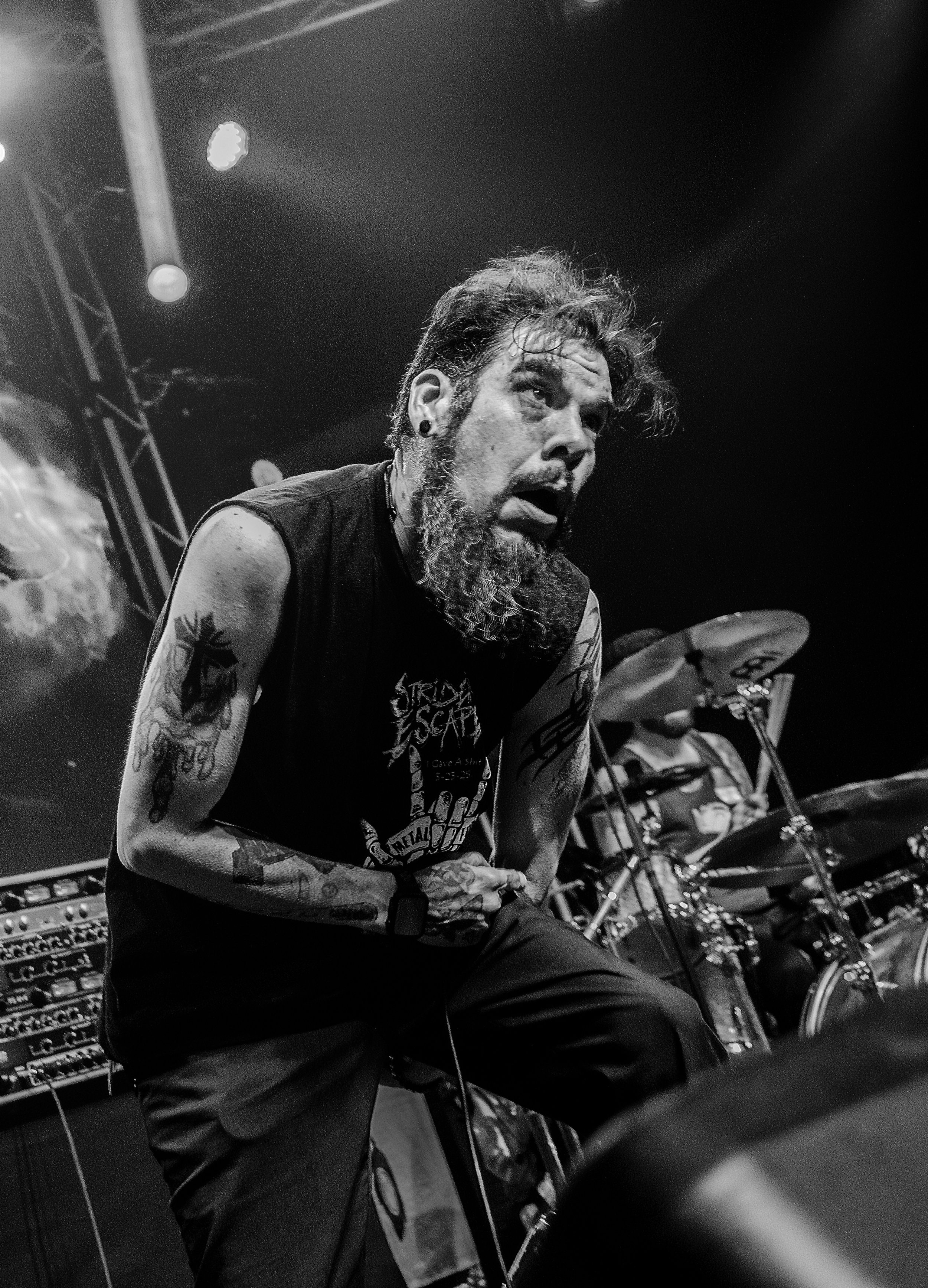 Black and white photo of a male musician with long hair and a beard, performing under stage lights, surrounded by musical equipment including drums and amplifiers.