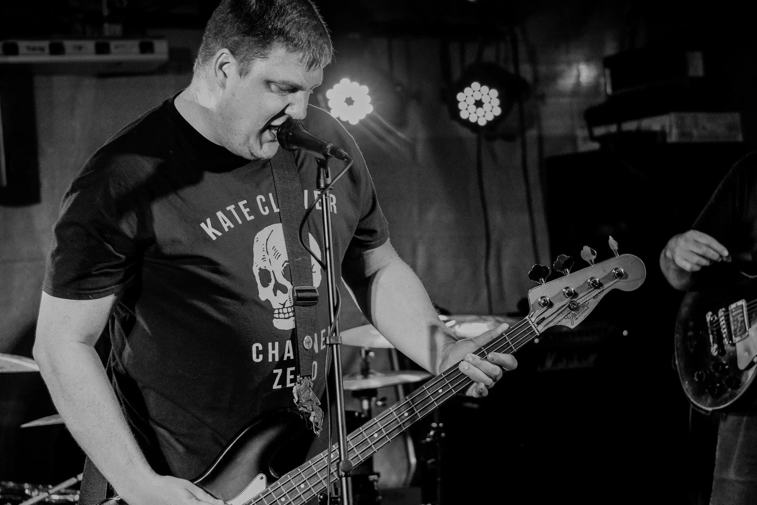 A young man performs on stage with a guitar in a black-and-white photo. He is singing into a microphone, wearing a T-shirt with a skull graphic and text. Stage lights and a keyboard are visible in the background.