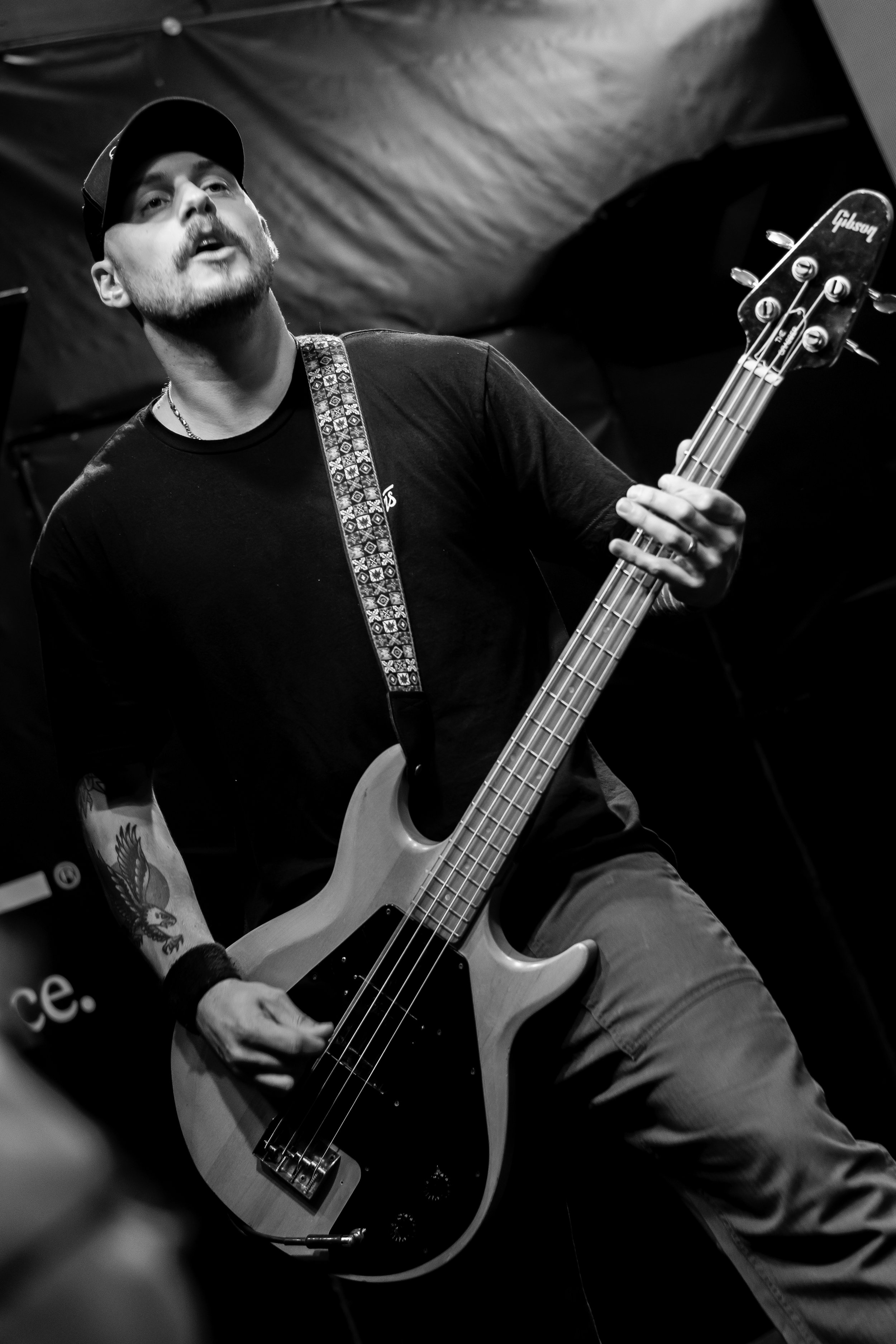 A man with a beard and tattoos playing an electric bass guitar on stage, wearing a baseball cap and a black t-shirt, in black and white photo.