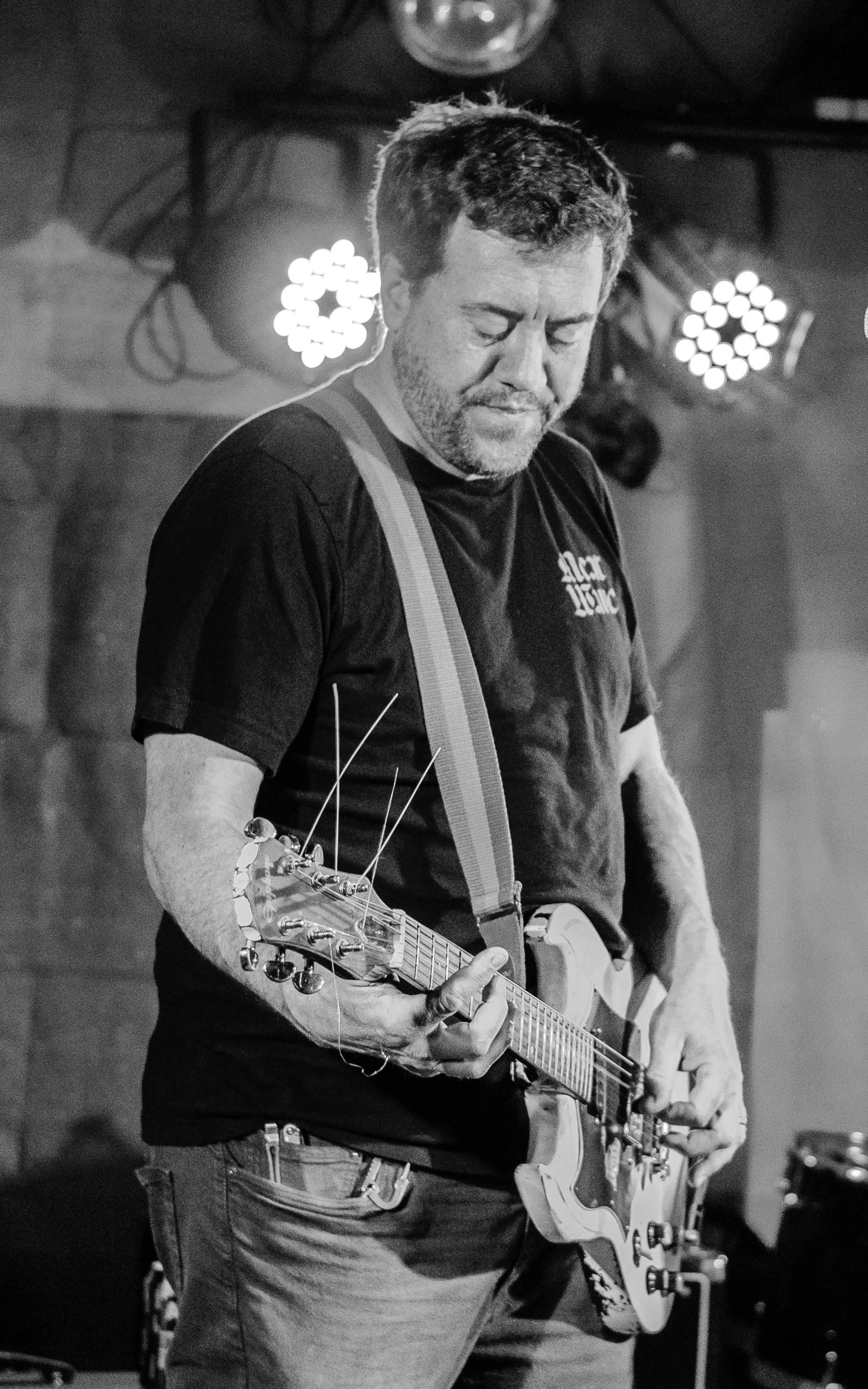 A man with short hair and a beard is playing an electric guitar. He is wearing a dark t-shirt and has a strap over his shoulder. He appears focused on playing, with his eyes closed or looking down. The background includes stage lighting and equipment
