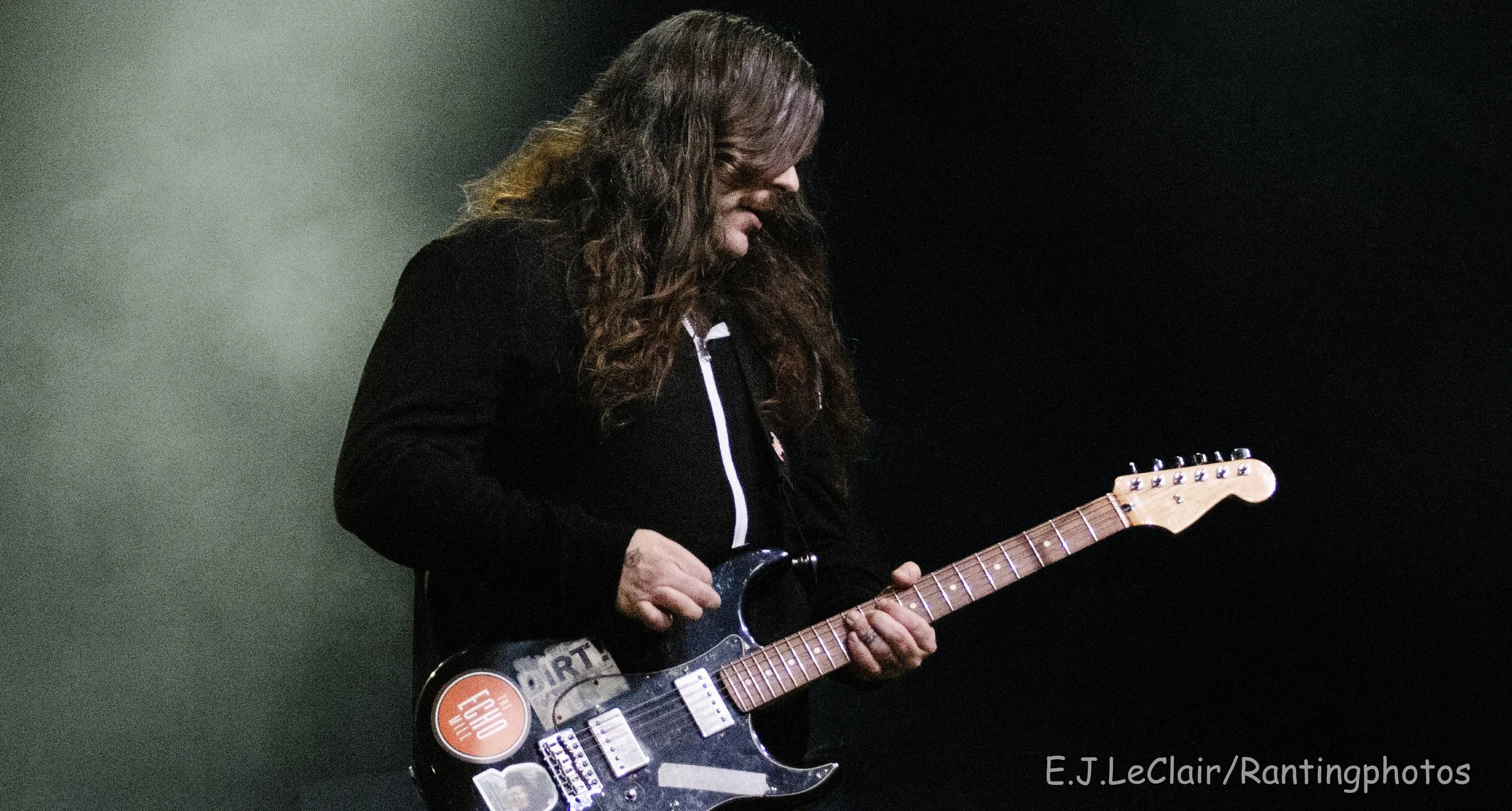 A man with long hair playing an electric guitar with stickers on it, standing against a dark background.