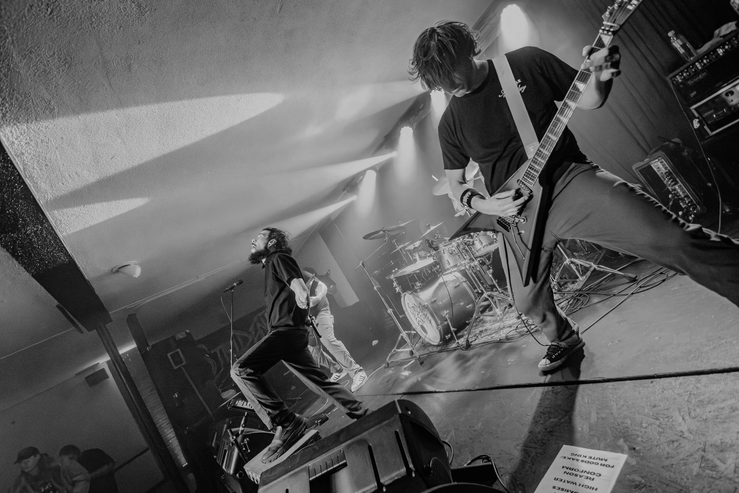 Two musicians perform on stage with one playing guitar and singing into a microphone, and the other playing drums in the background, under stage lights.