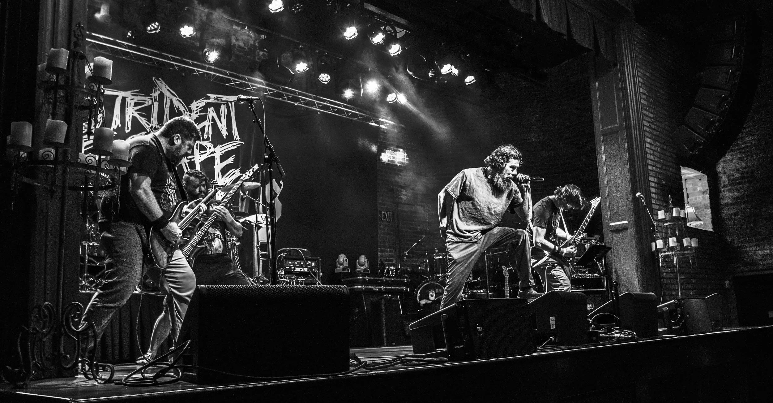 A black and white photo of a band performing on stage at a music venue. Four members are visible: a vocalist kneeling and singing into a microphone, and three guitarists playing their instruments. The stage has lighting and sound equipment, with a la
