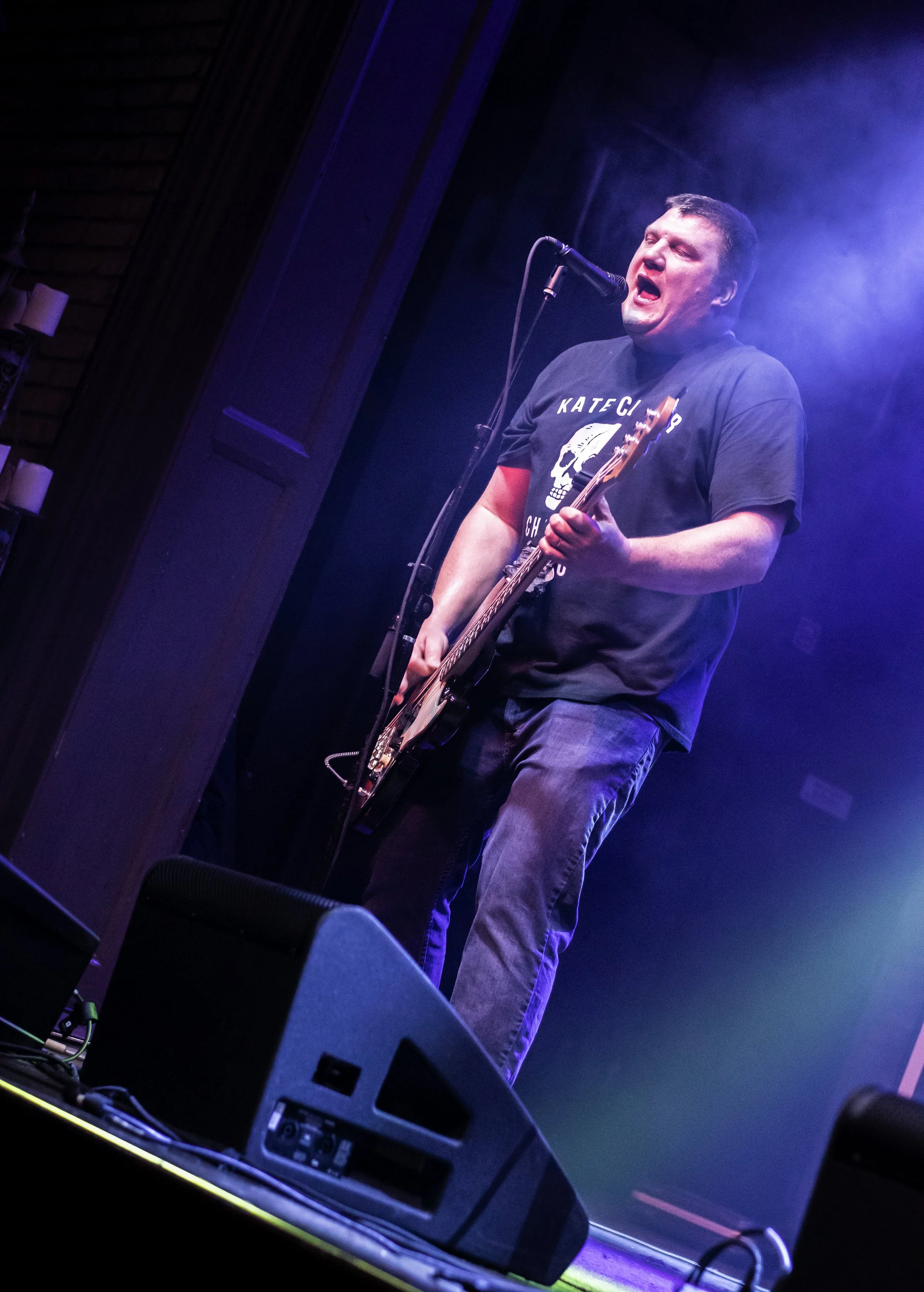 A man performing on stage, singing into a microphone and playing an electric guitar, with stage lights creating a misty atmosphere