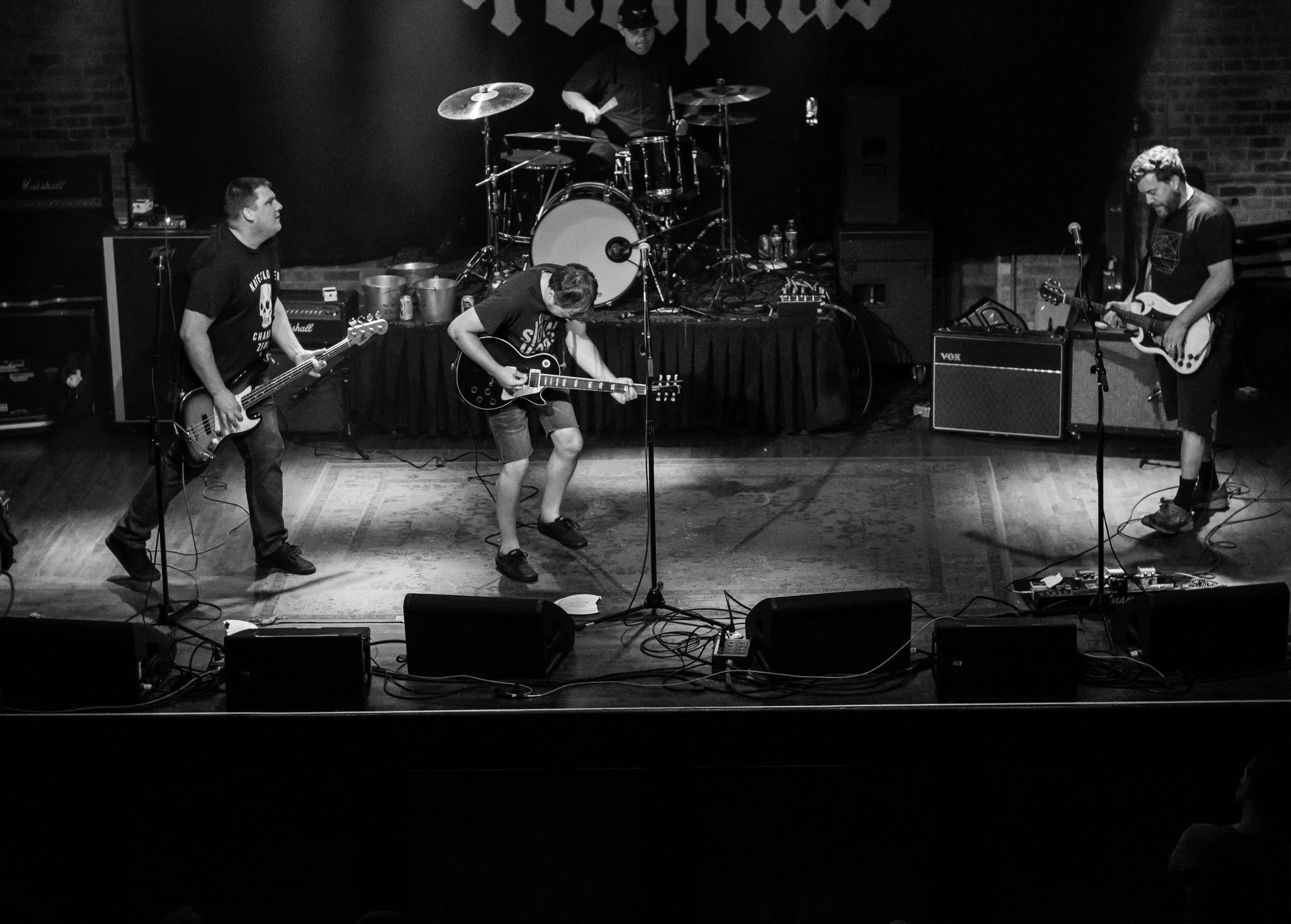 Black and white photo of a four-member band performing on stage with musical instruments, including electric guitars, drums, and amplifiers.