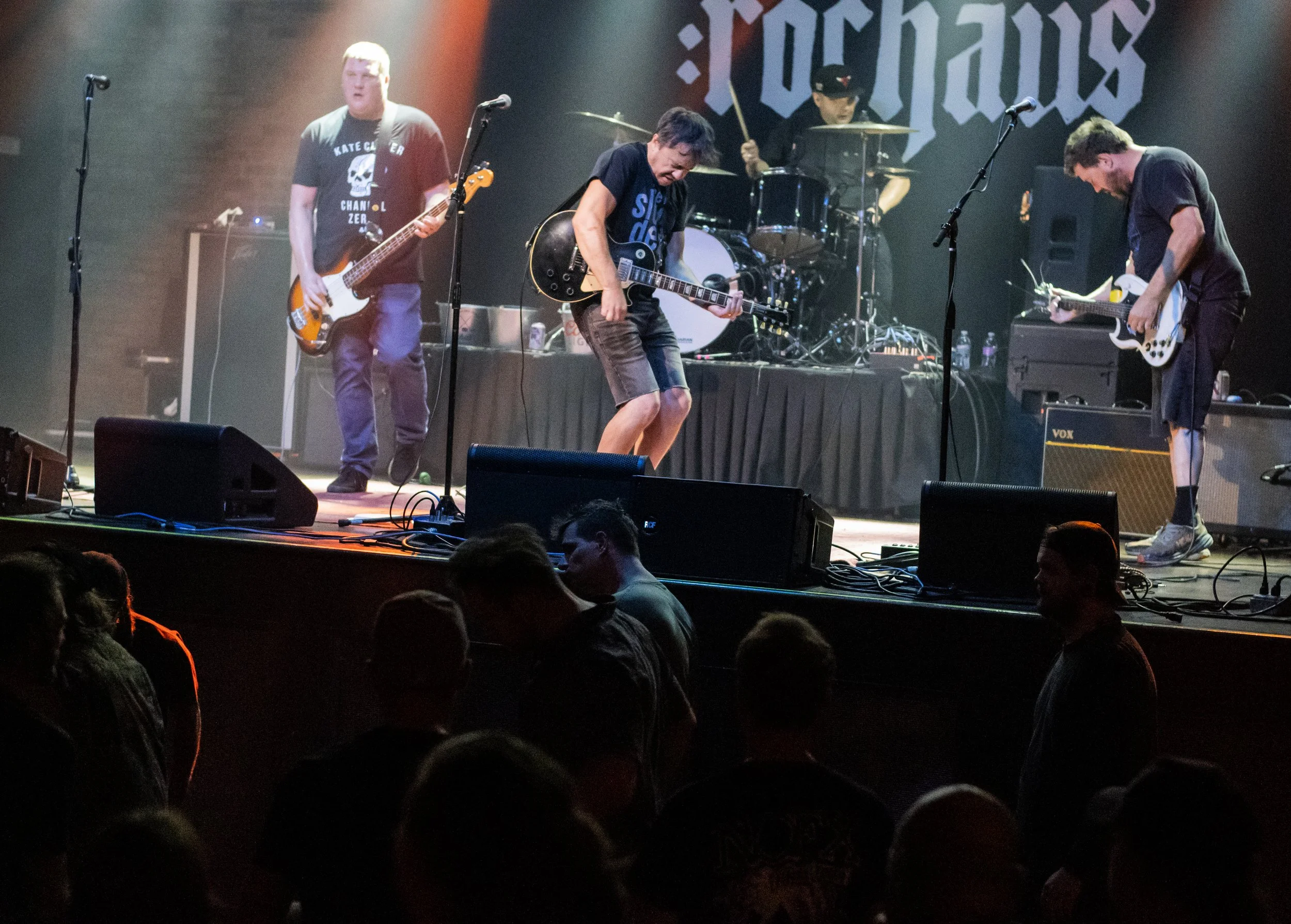 A band performs on stage with four members, including guitarists and a drummer, during a concert. Audience members are visible in the foreground.