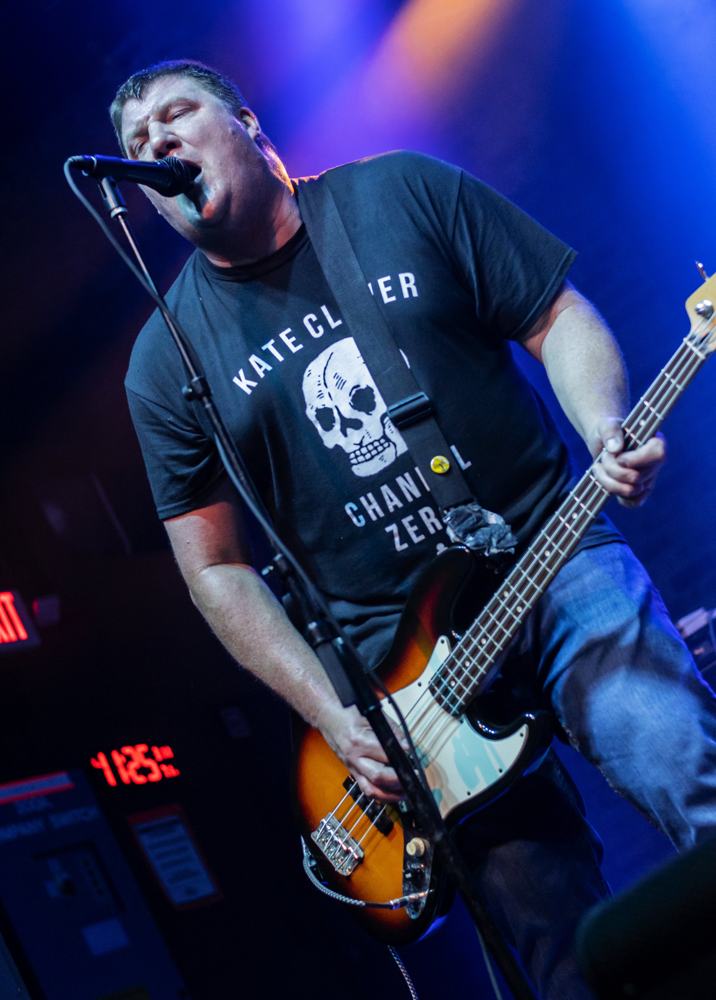 A man singing into a microphone while playing an electric bass guitar on stage, wearing a black shirt with a skull graphic and text, with colorful stage lighting.